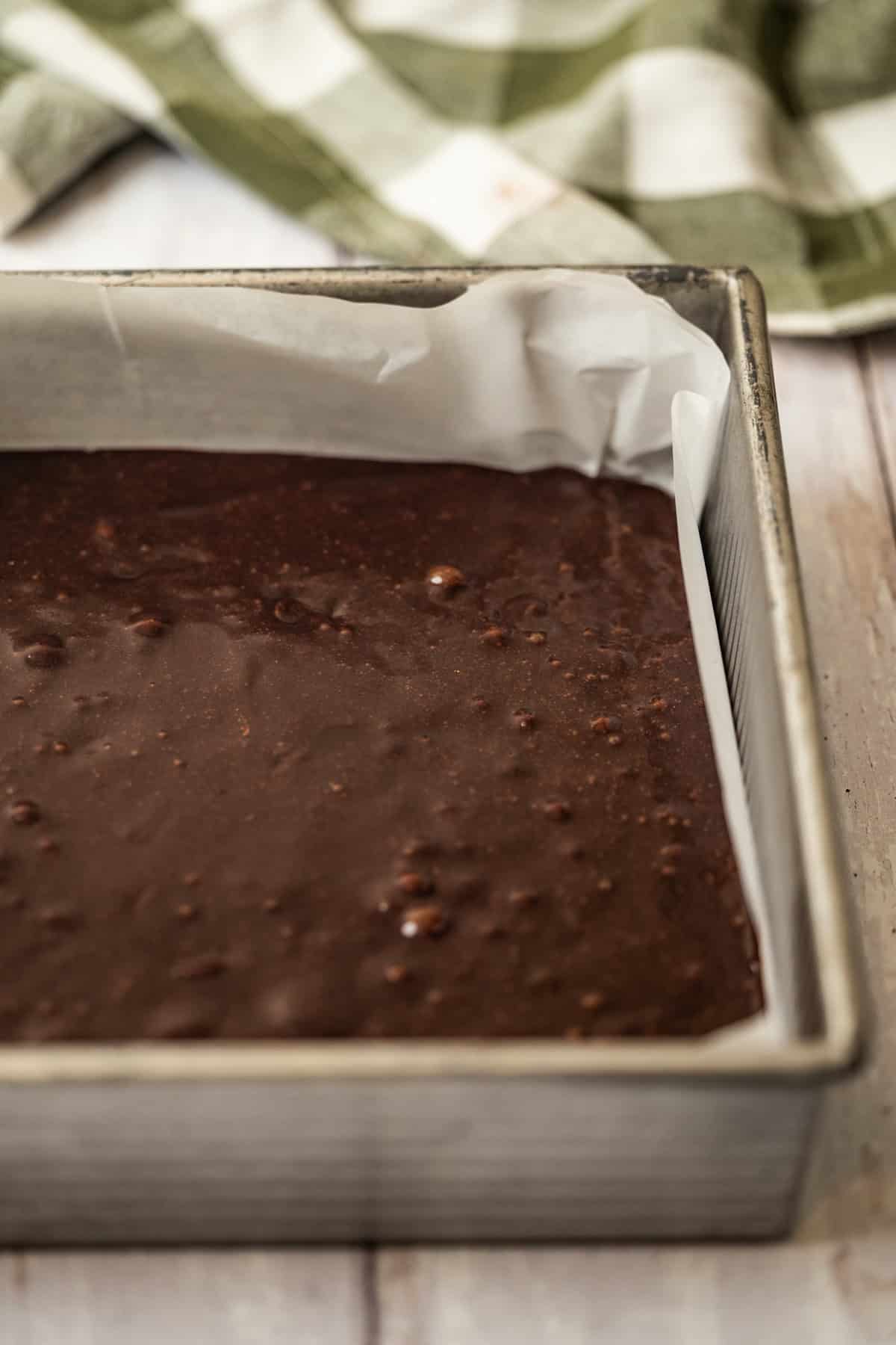 A square metal baking pan lined with parchment paper is filled with chocolate brownie batter, ready to be baked. A green and white checkered cloth is in the background on a light wooden surface.
