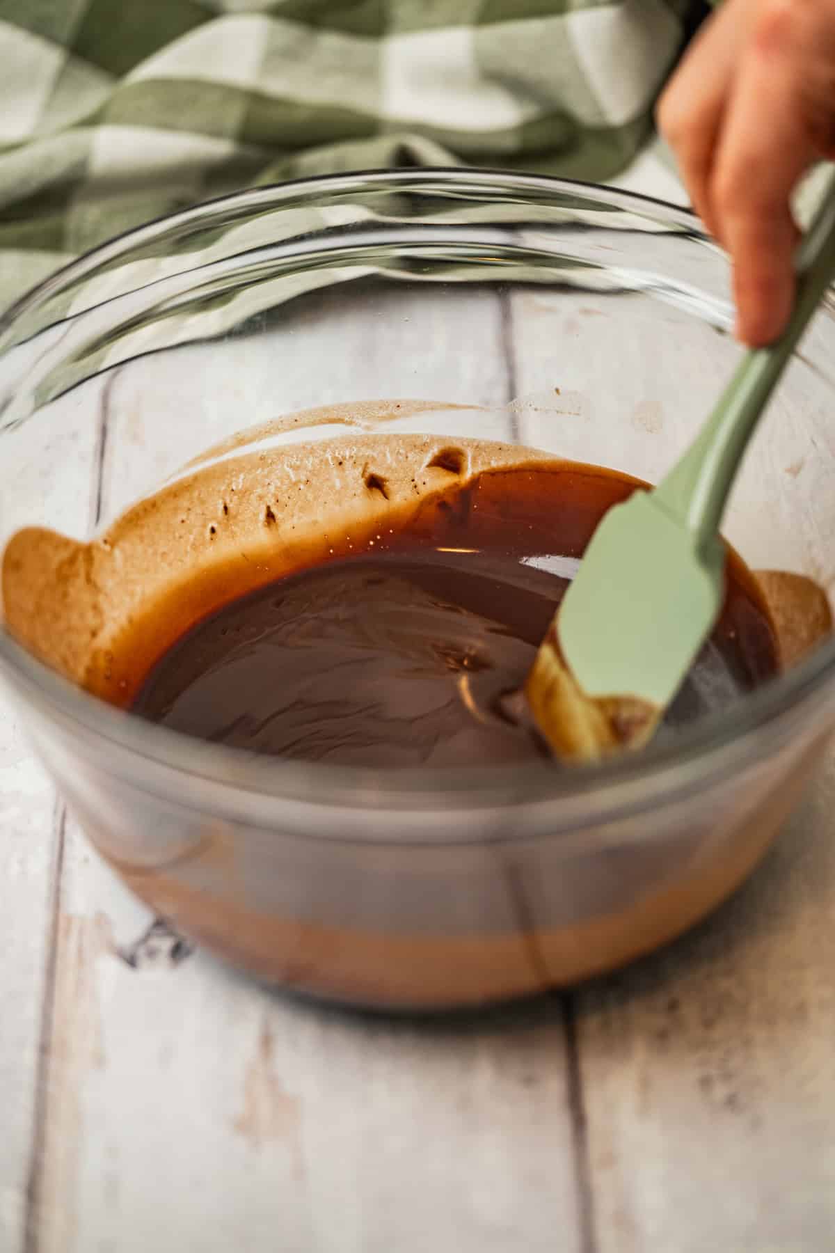 A close-up of a person stirring melted chocolate in a clear glass bowl with a green spatula on a light wooden surface; a green and white cloth is in the background.