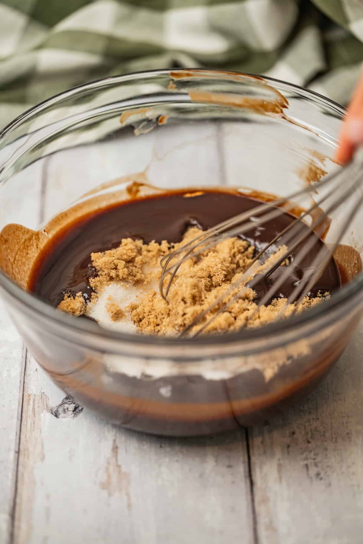 A hand whisks brown sugar into a bowl of melted chocolate mixture on a light wooden surface, with a green checkered cloth in the background.