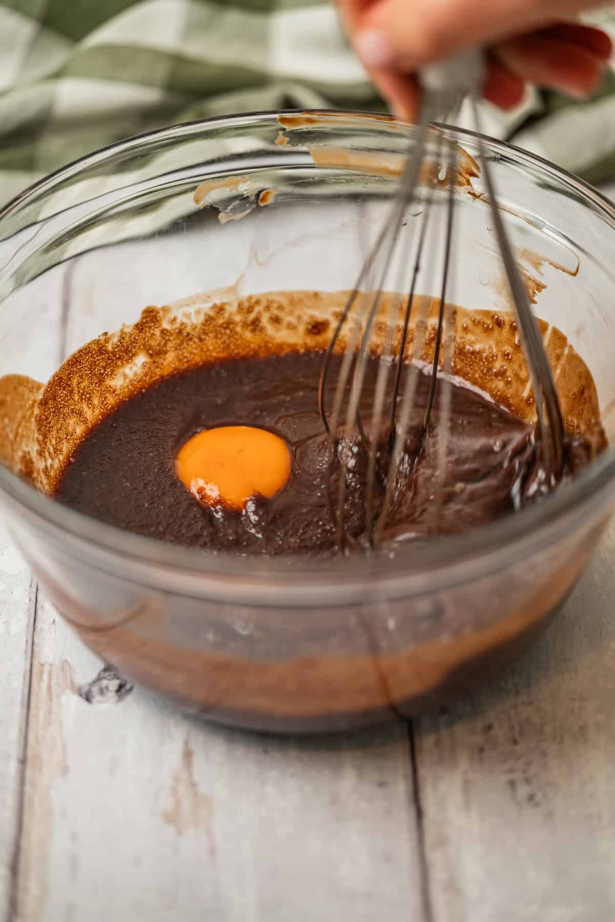 A hand uses a whisk to mix a raw egg into a bowl of chocolate batter on a light wooden surface.