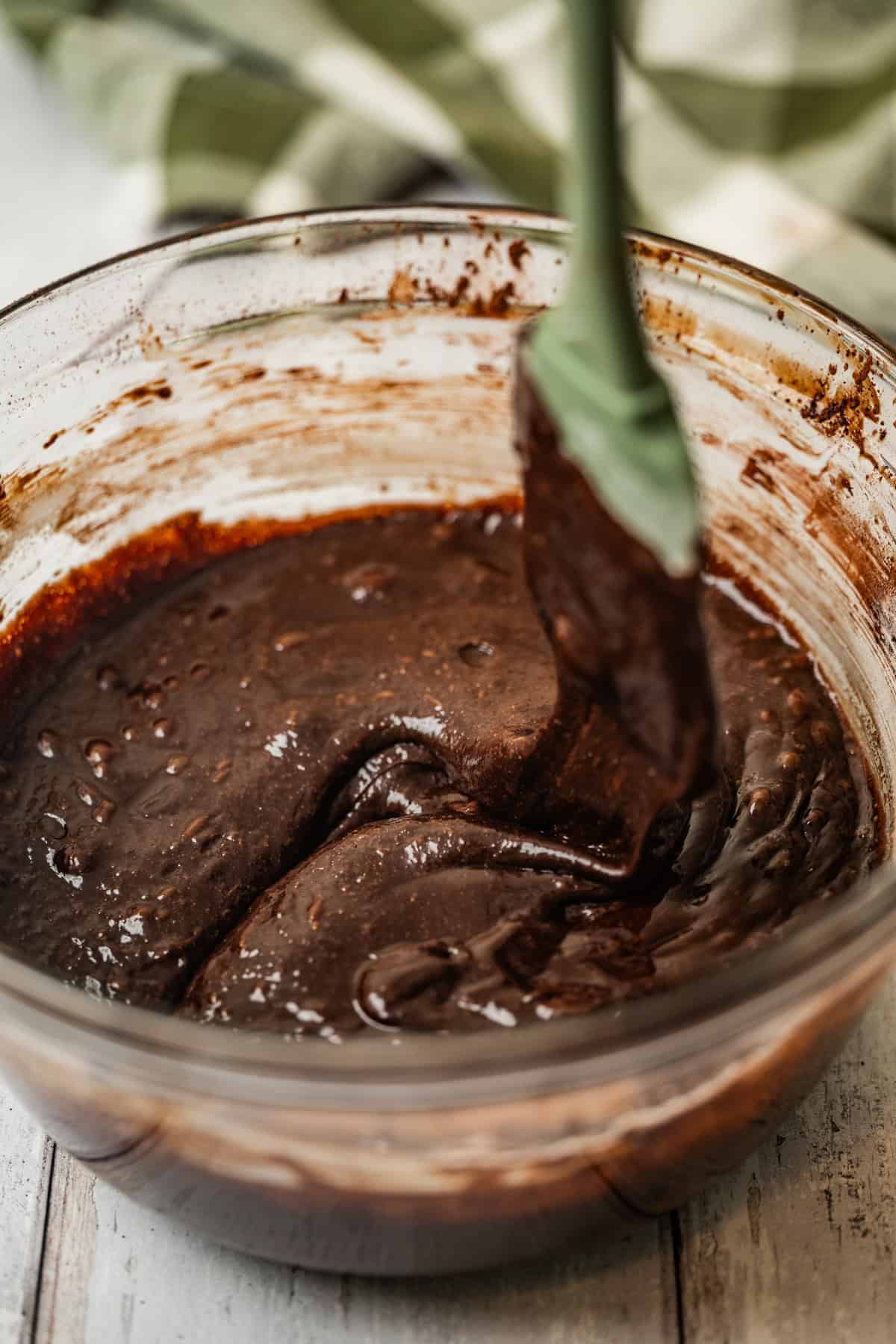 A glass bowl filled with chocolate brownie batter being mixed with a green spatula, on a white wooden surface. A green and white checkered cloth is visible in the background.