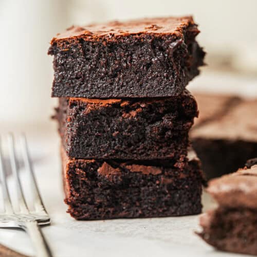 Three thick, fudgy chocolate brownies are stacked on top of each other on a piece of parchment paper, with two forks beside them and more brownies in the blurred background.