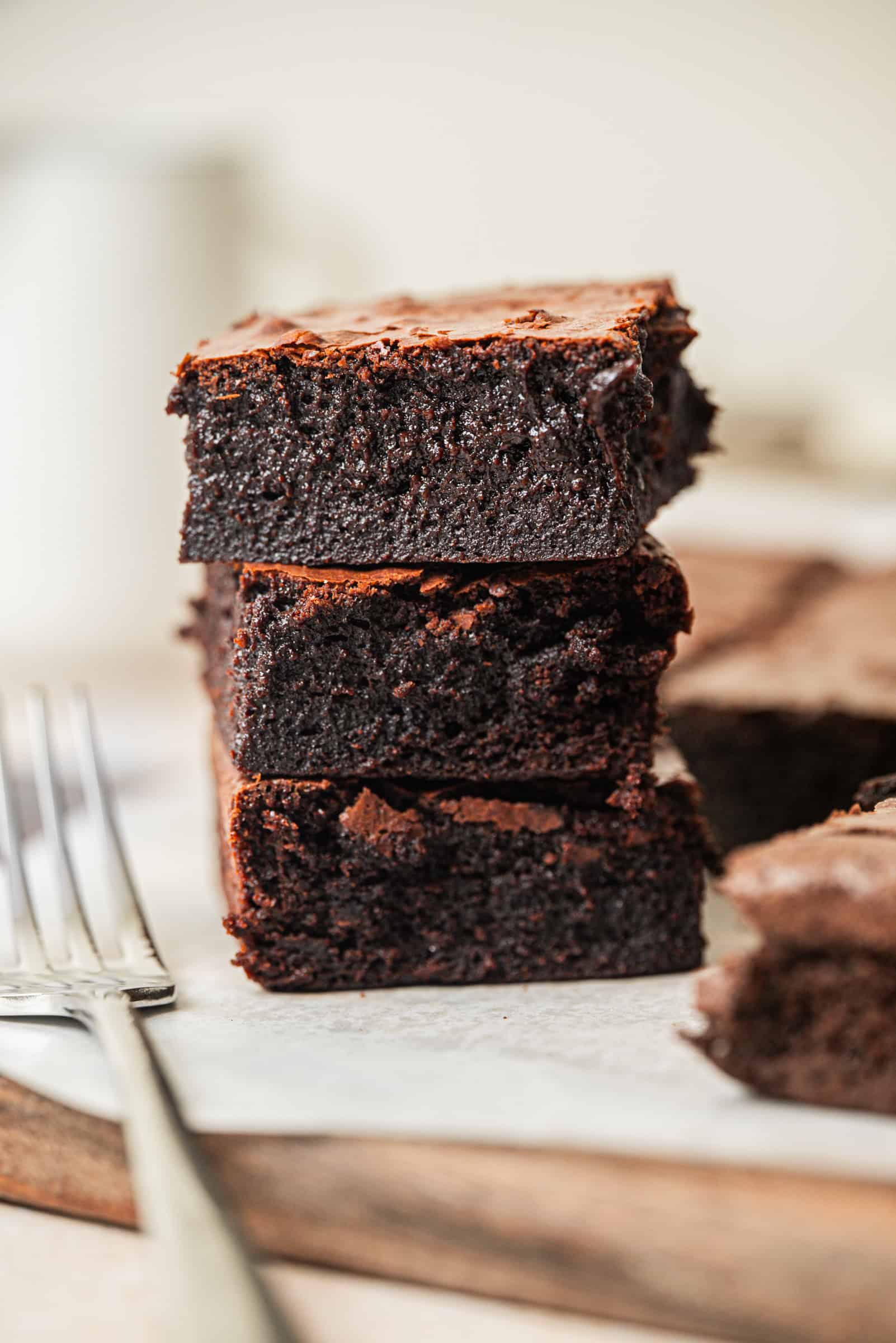 Three thick, fudgy chocolate brownies are stacked on top of each other on a piece of parchment paper, with two forks beside them and more brownies in the blurred background.