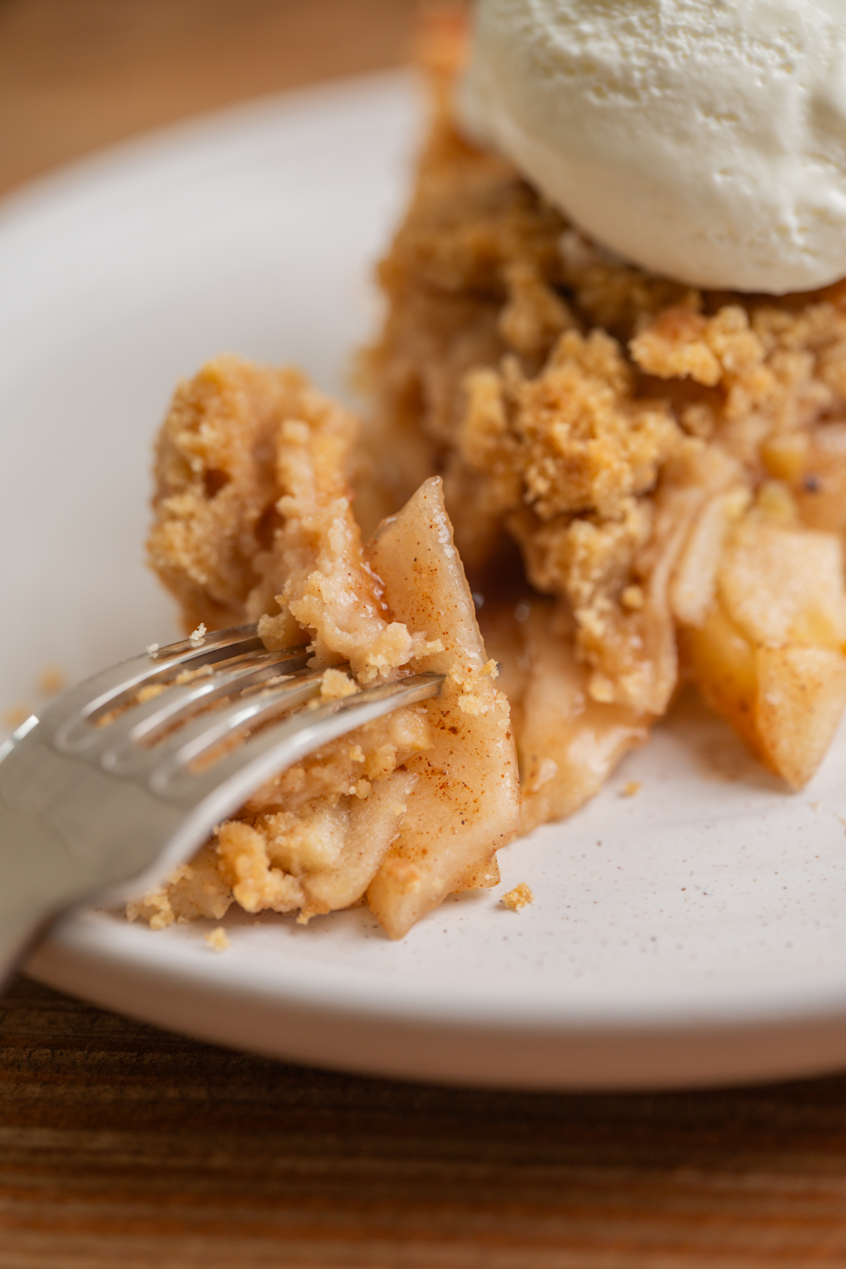 A close-up of a fork cutting into a slice of apple crumble pie topped with a scoop of vanilla ice cream on a white plate.