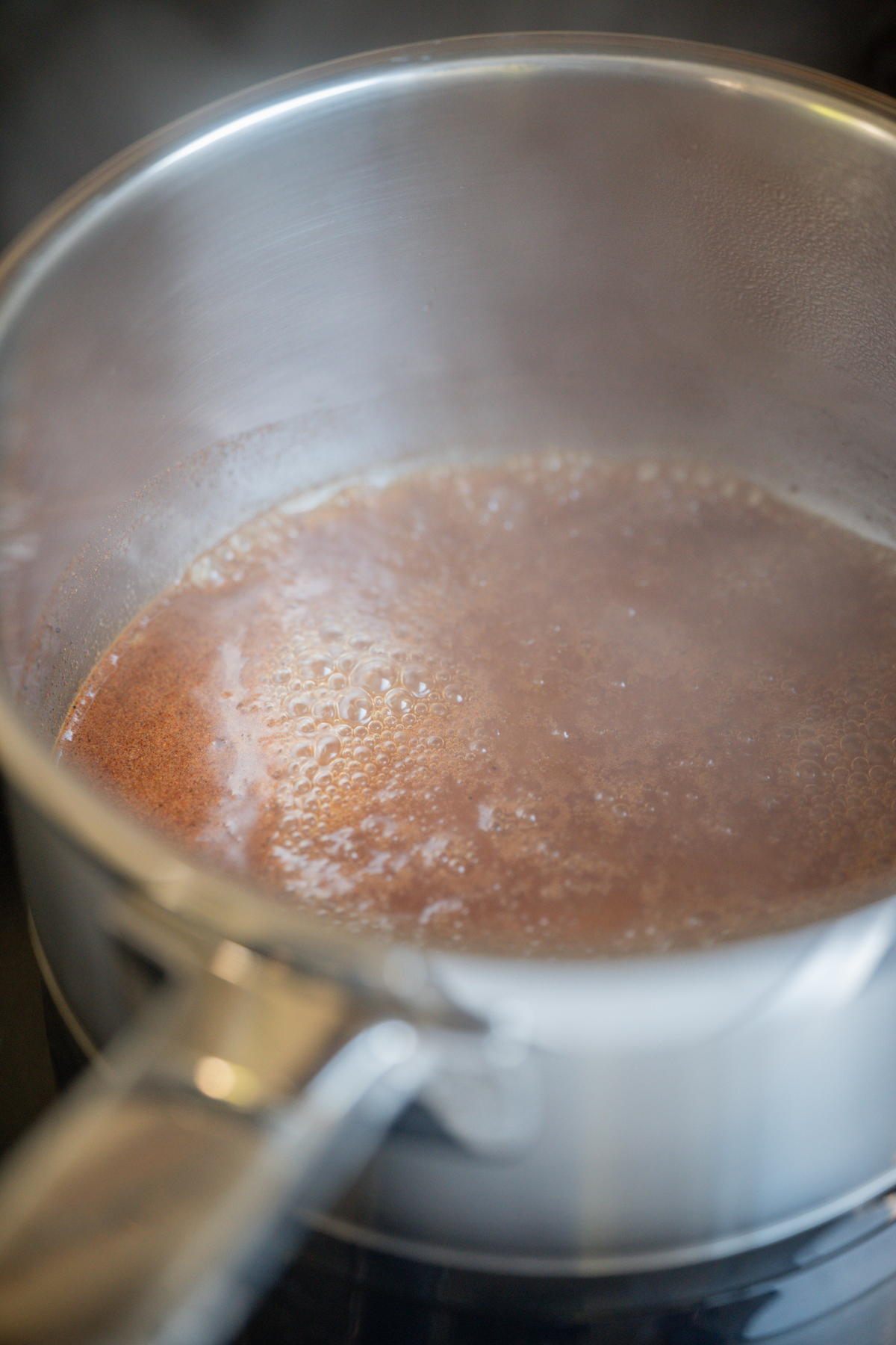 A close-up of a stainless steel saucepan with a brown liquid simmering and small bubbles forming on the surface, with steam rising from the pot.