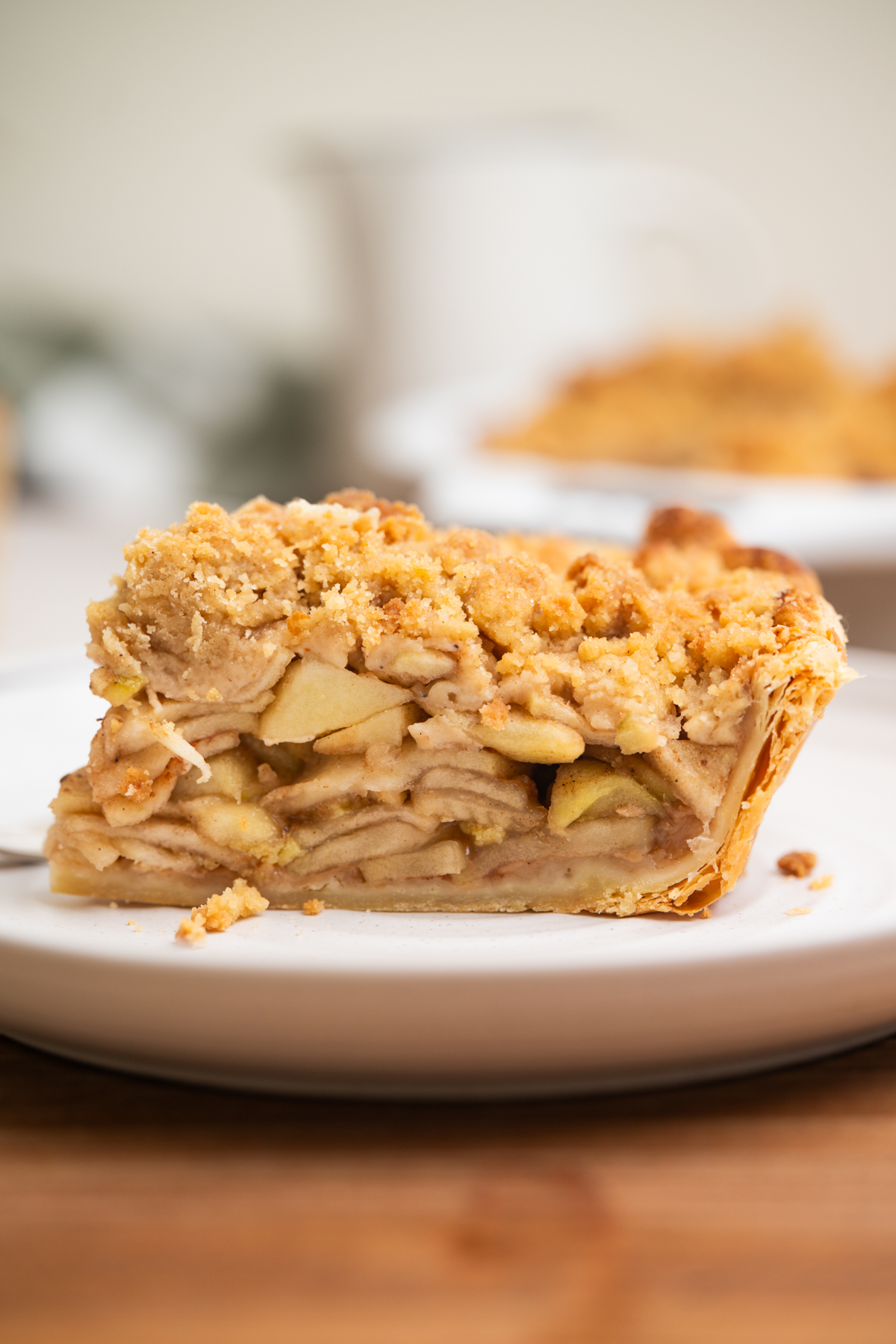 A close-up of a slice of apple crumble pie on a white plate, showing layers of sliced apples and a crumbly topping. The background is softly blurred.