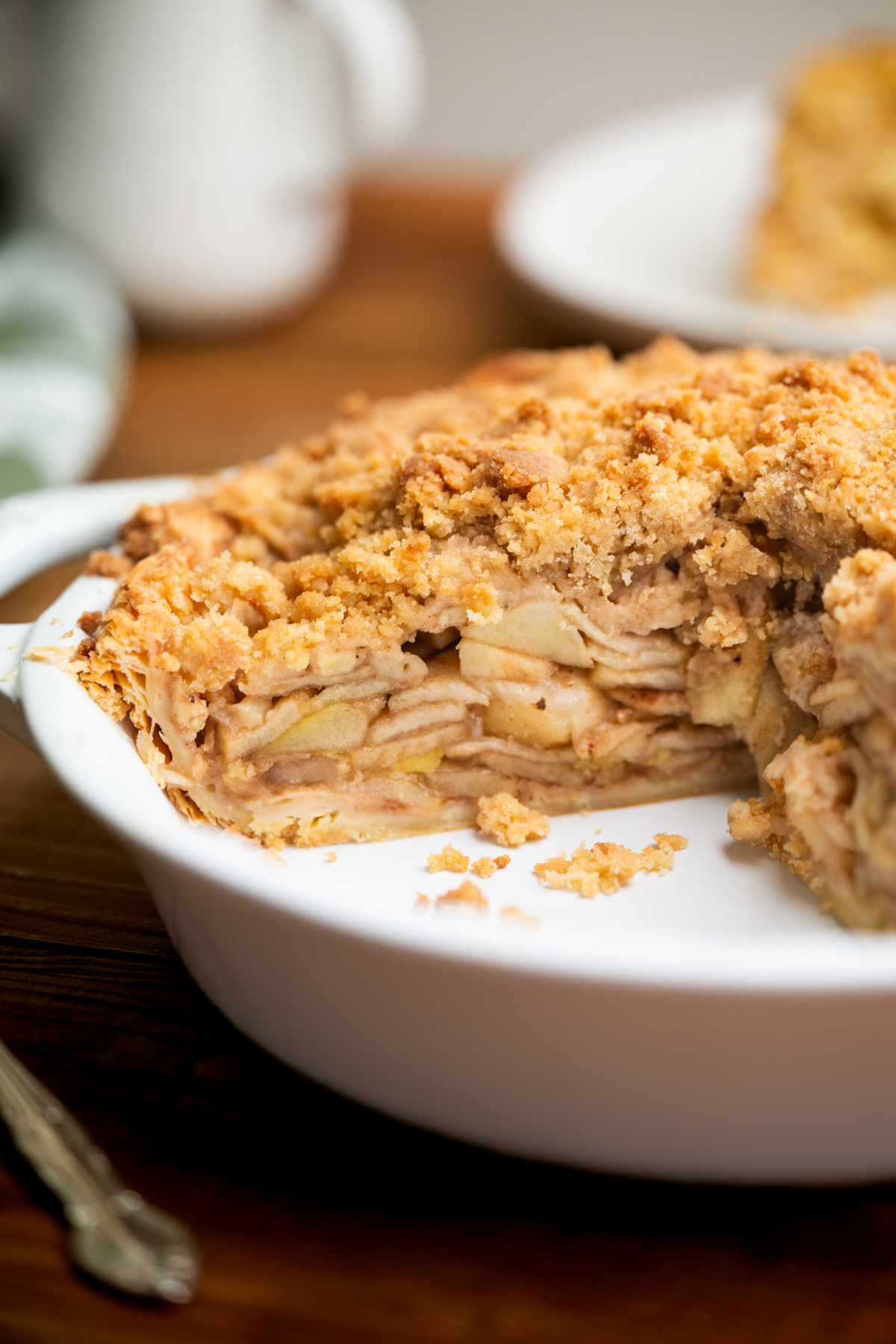 A close-up of a sliced apple crumble pie in a white pie dish, showing layers of apple filling and a crumbly golden topping, with crumbs scattered on the dish.