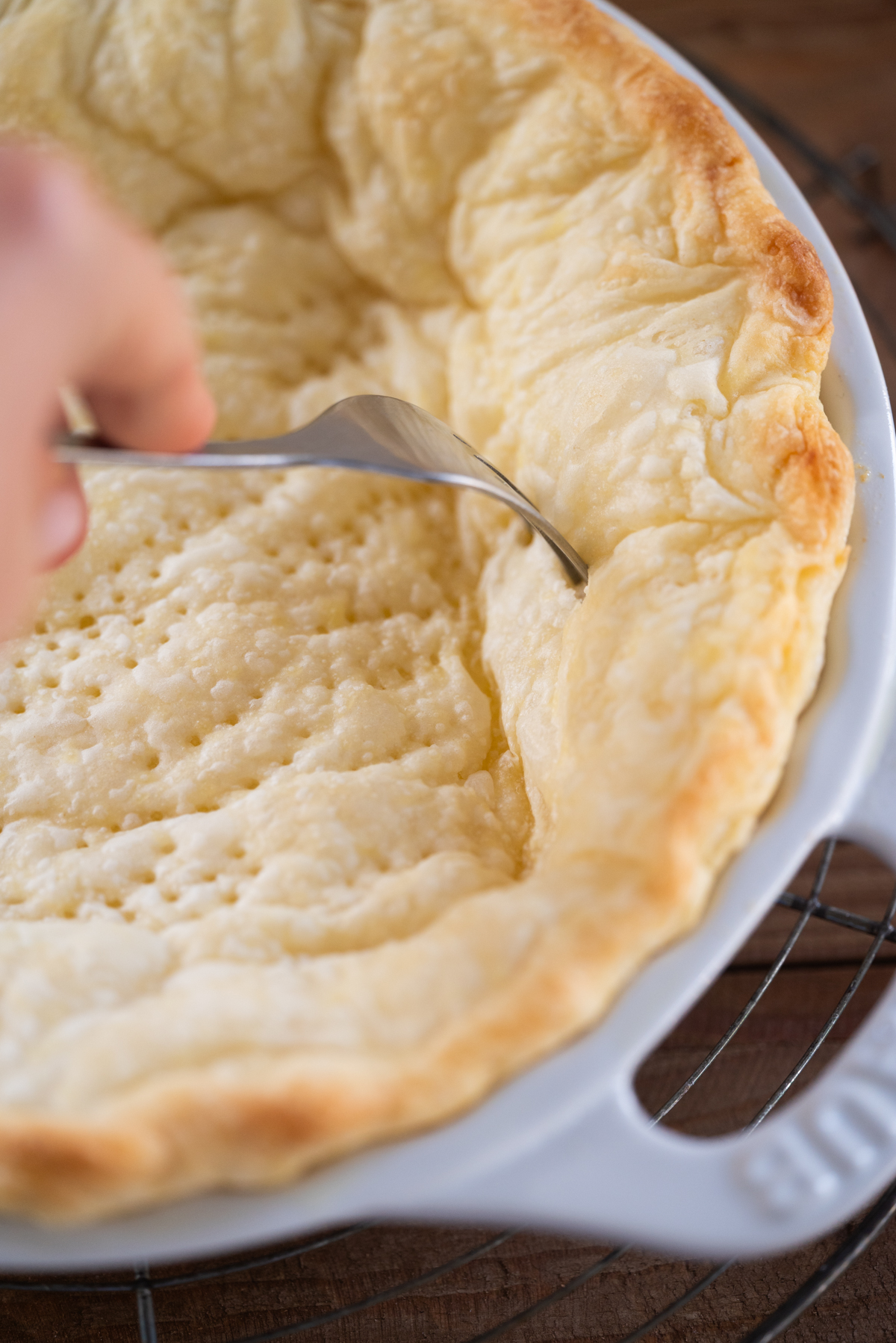 A hand uses a fork to poke holes in a baked, golden pie crust in a white dish, making small perforations in the bottom and sides.