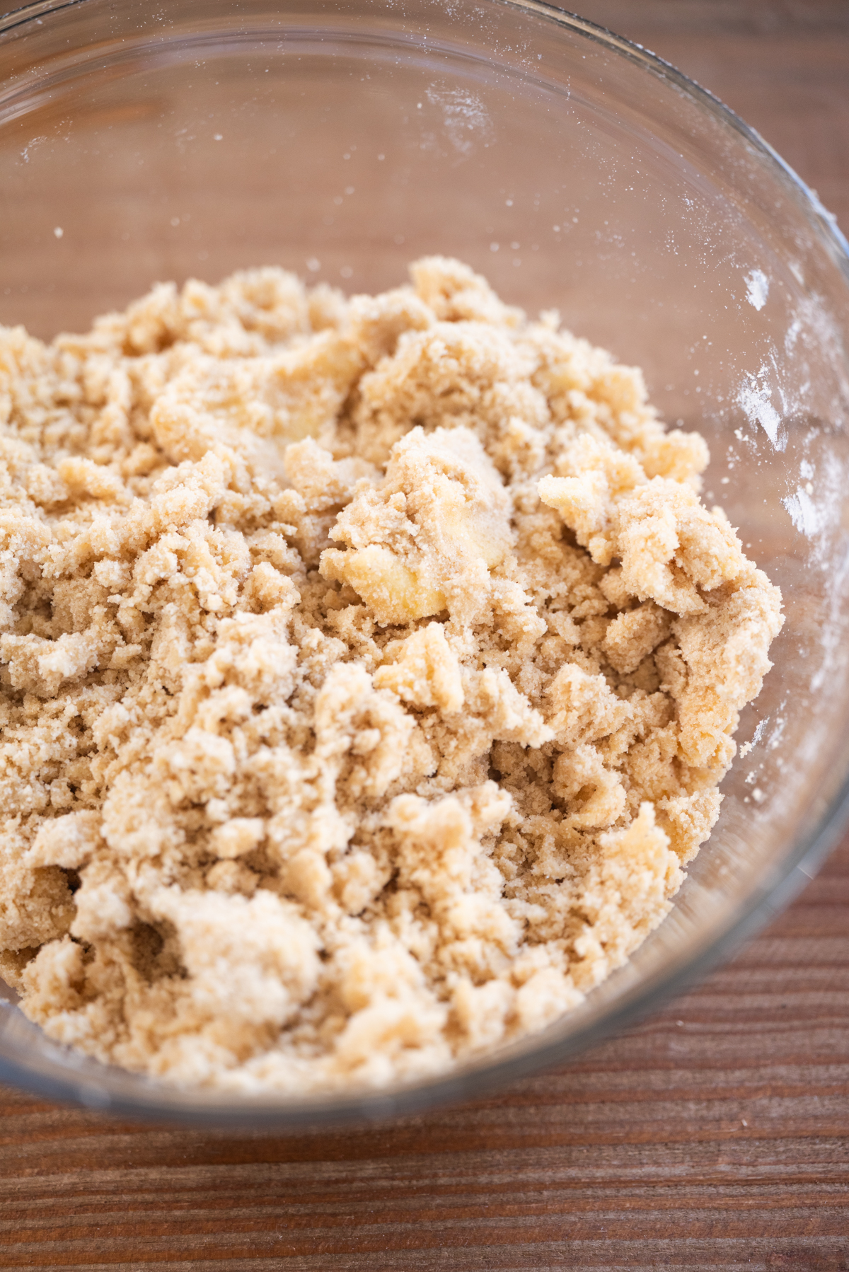 A clear glass bowl filled with crumbly, pale yellow dough mixture sits on a wooden surface.