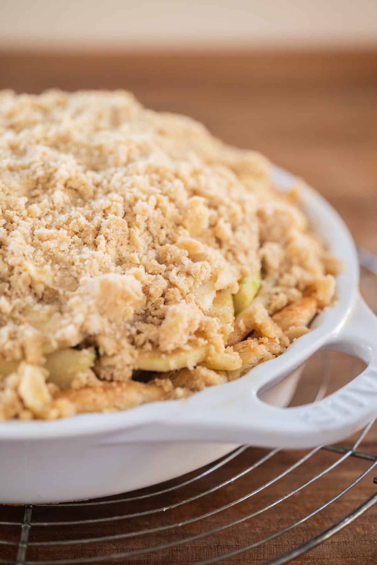 A close-up of a freshly baked apple crumble pie in a white ceramic pie dish, sitting on a round cooling rack, with a golden, crumbly topping and visible apple slices.