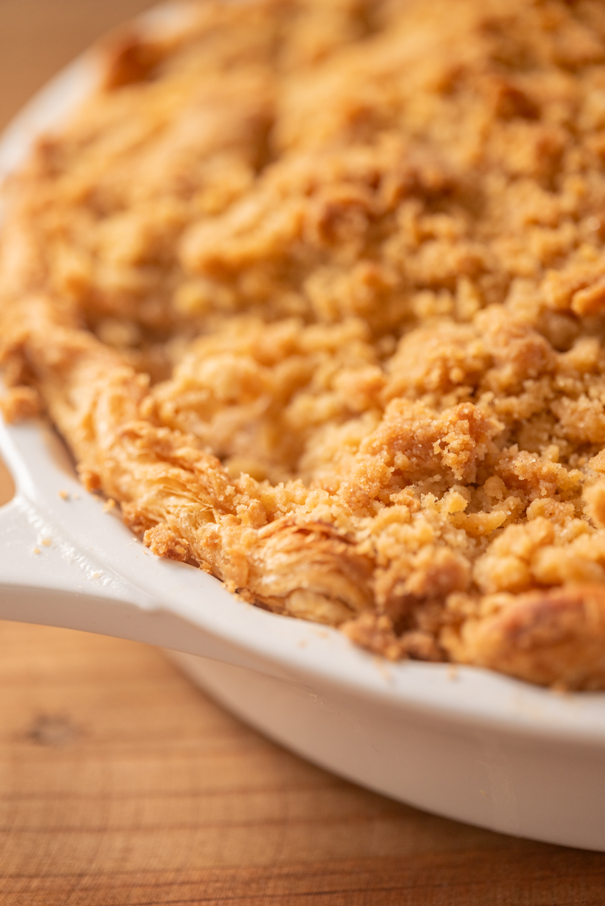 Close-up of a golden, crumbly pie with a flaky crust in a white ceramic dish, sitting on a wooden surface.