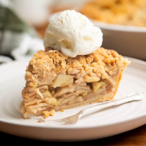 A slice of apple crumb pie topped with a scoop of vanilla ice cream sits on a white plate with a fork, with a blurred pie and mug in the background.