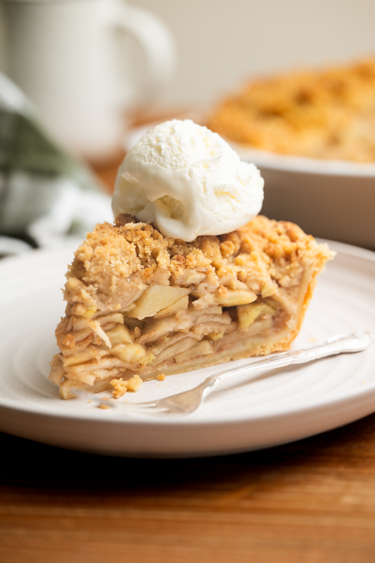 A slice of apple crumb pie topped with a scoop of vanilla ice cream sits on a white plate with a fork, with a blurred pie and mug in the background.
