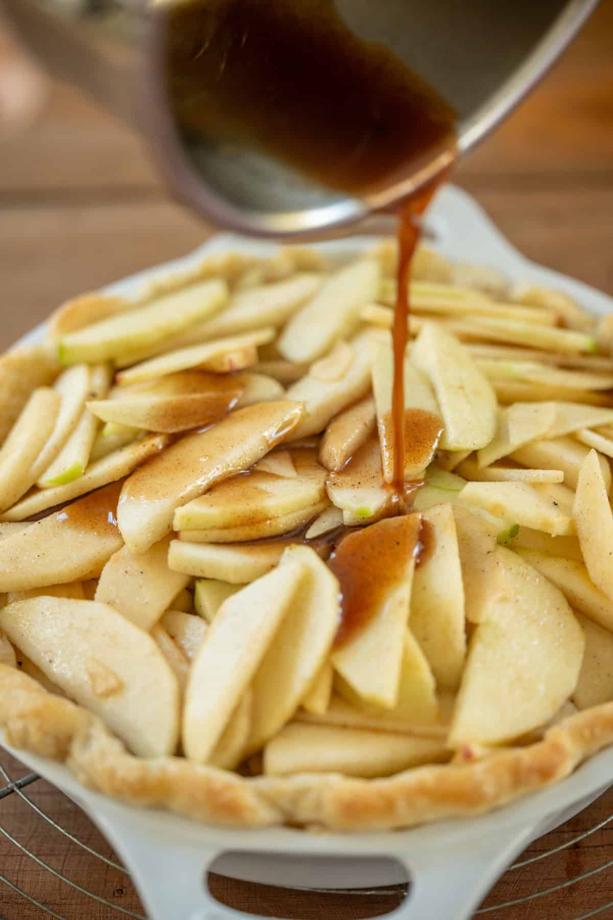 A close-up of sliced apples arranged in a pie crust as a brown sauce is being poured over the apples from a saucepan.