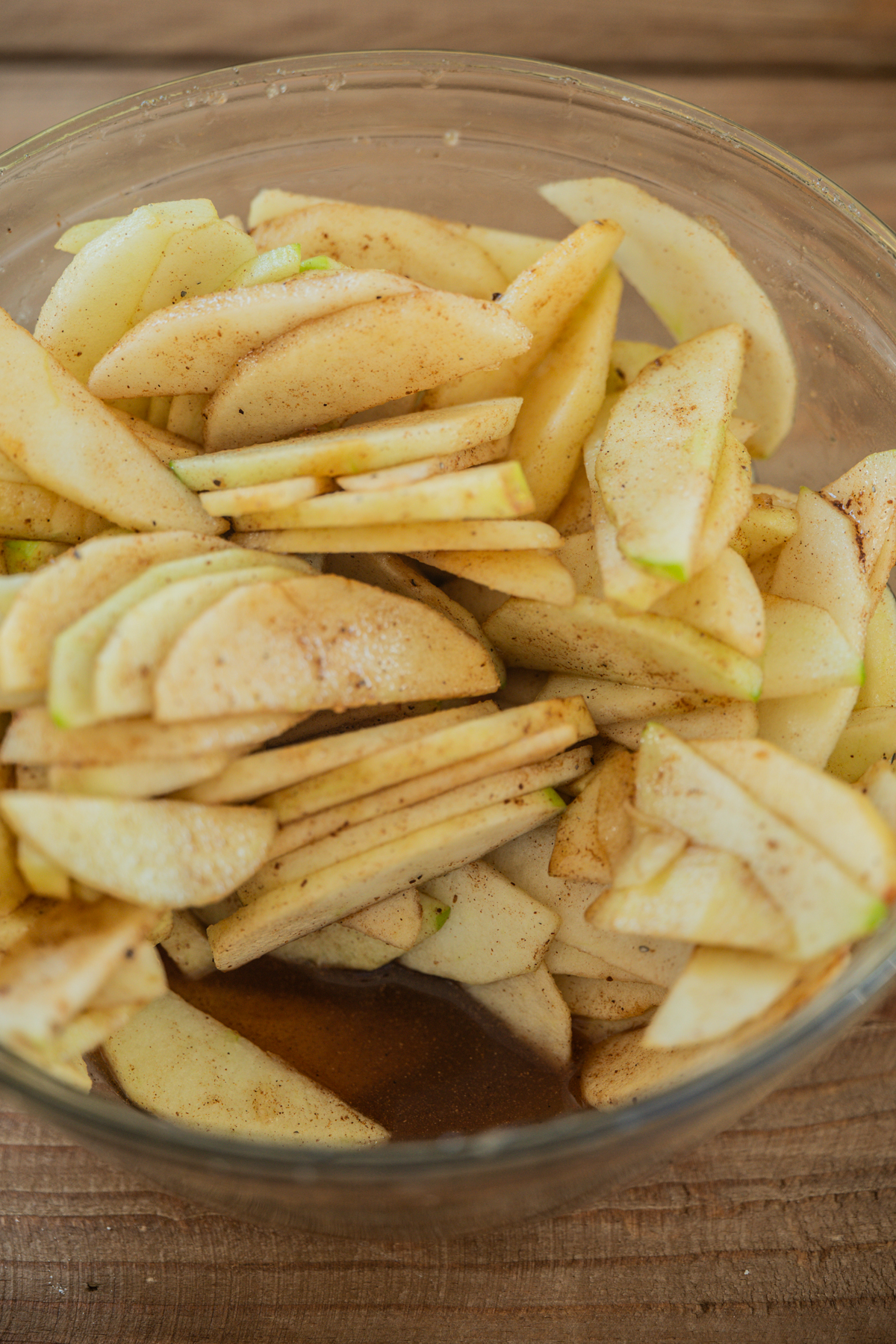 A glass bowl filled with thinly sliced apples coated in cinnamon and sugar, sitting on a wooden surface.