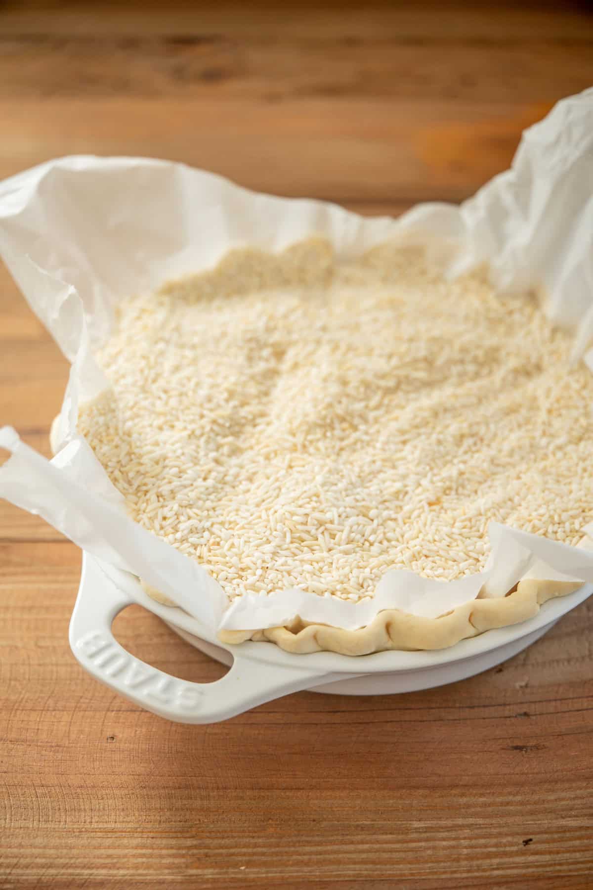 A pie crust lined with parchment paper and filled with uncooked rice, used as pie weights, sits in a white ceramic dish on a wooden surface.