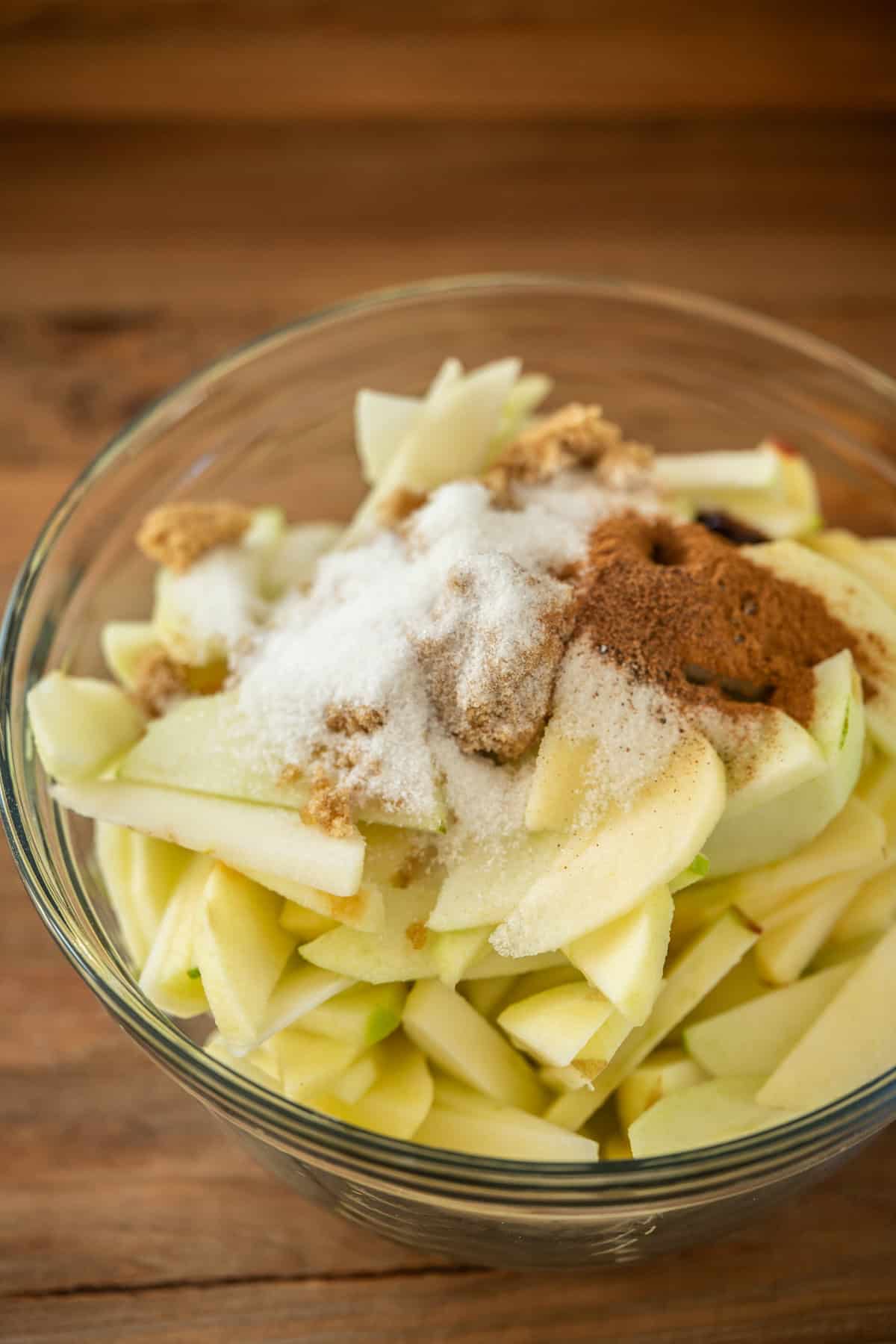 A glass bowl filled with sliced apples, topped with granulated sugar, brown sugar, and ground cinnamon, sits on a wooden surface.