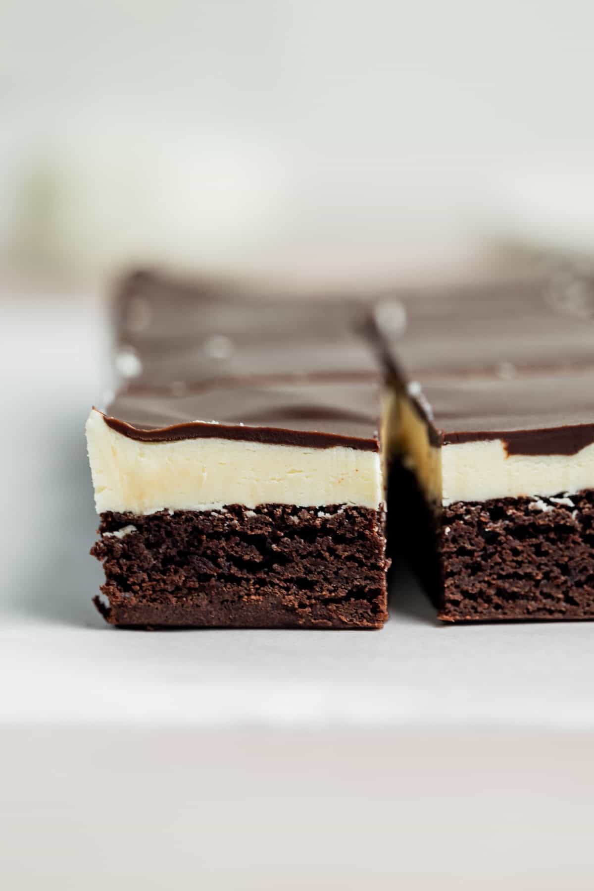 A close-up of a dessert bar with three layers: a dense chocolate base, a creamy white middle layer, and a glossy chocolate topping, neatly cut into squares.