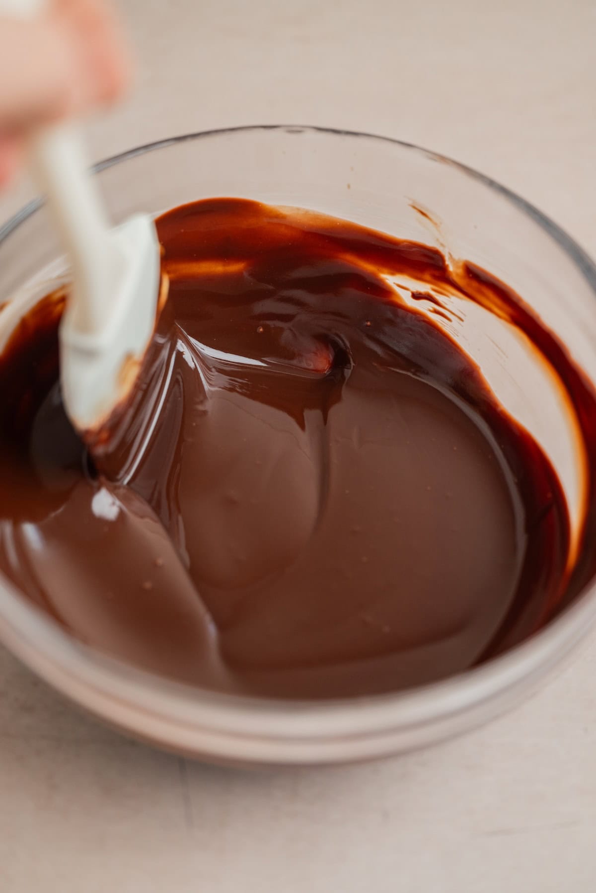 A spatula stirs smooth, glossy melted chocolate in a clear glass bowl on a light-colored surface.