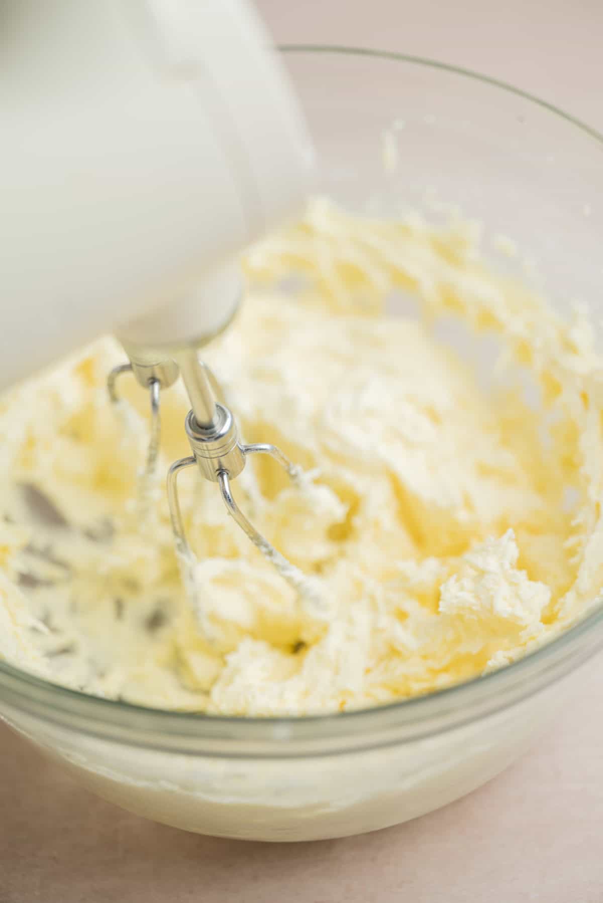 A close-up of an electric mixer blending a creamy, pale yellow mixture in a clear glass bowl, likely butter and sugar being creamed together for baking.