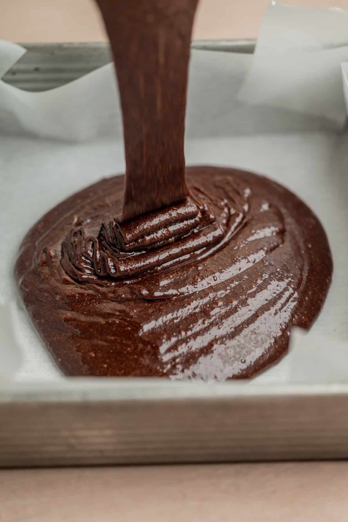 Thick chocolate brownie batter being poured into a parchment-lined baking pan, ready to be spread and baked.