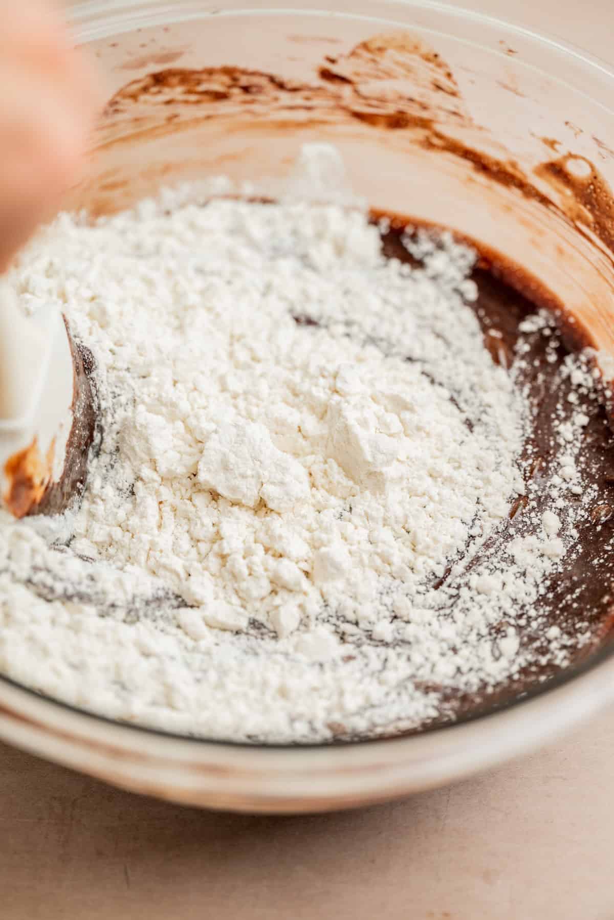 A close-up of a glass bowl with brownie batter being mixed. A spatula is stirring white flour into the dark chocolate mixture.