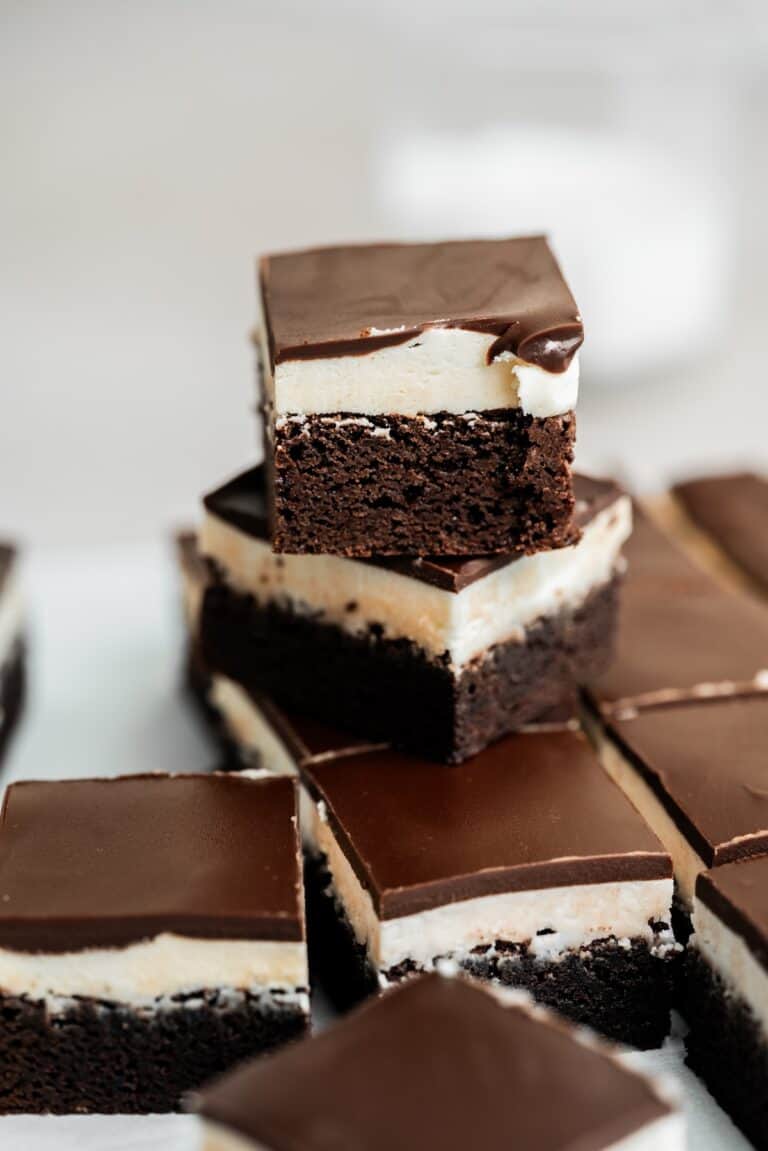 A close-up of chocolate brownies with a creamy white layer and glossy chocolate topping, stacked and arranged on parchment paper.