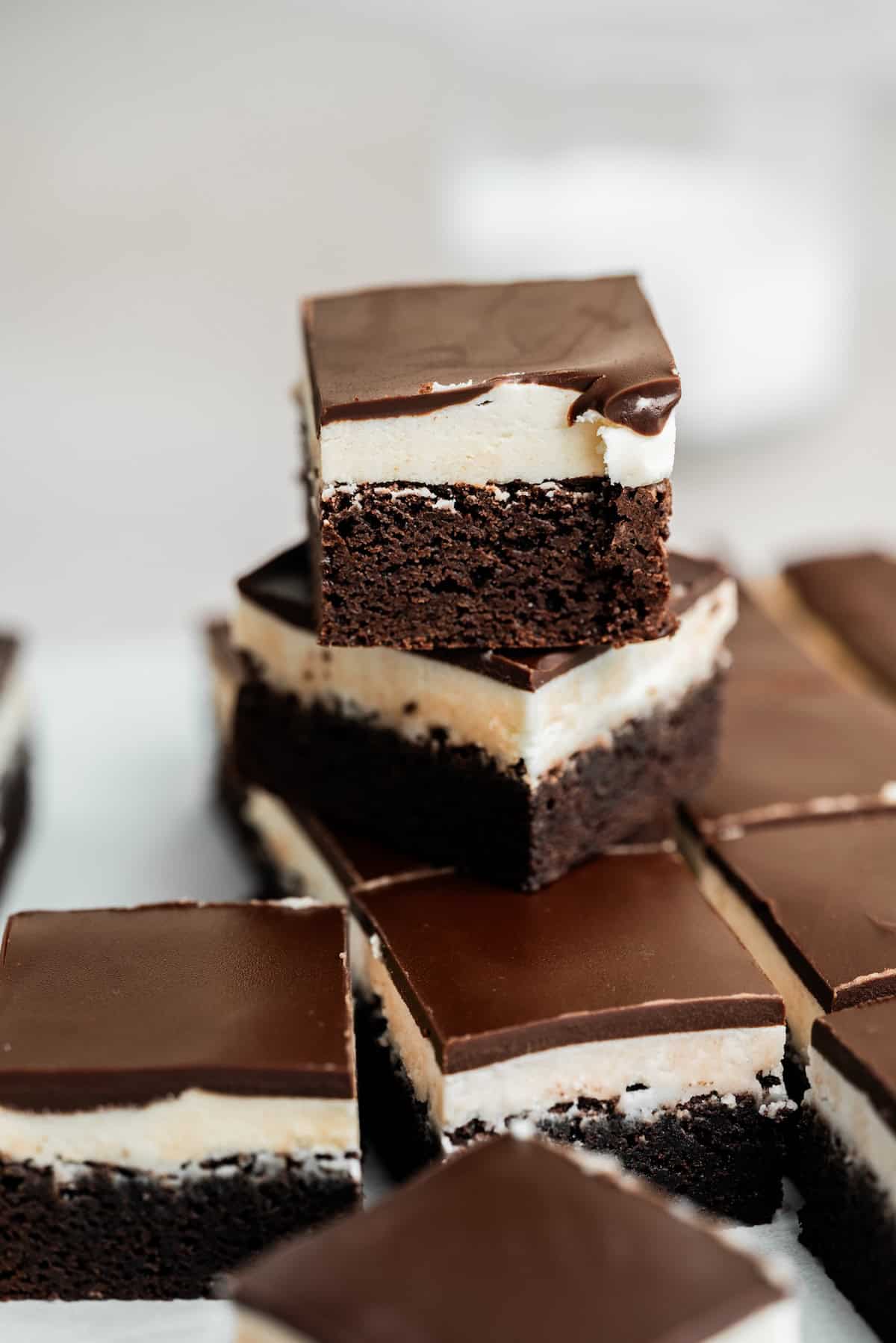 A close-up of chocolate brownies with a creamy white layer and glossy chocolate topping, stacked and arranged on parchment paper.