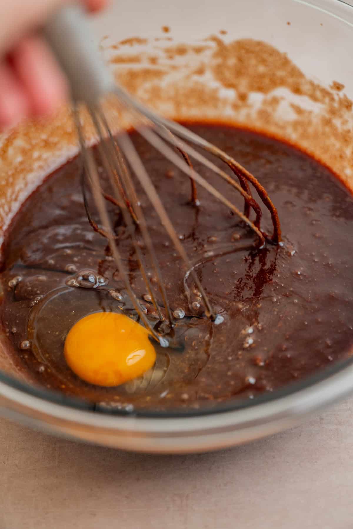 A close-up of a hand whisking a raw egg into a bowl of chocolate brownie batter. The egg yolk is clearly visible on top of the mixture.