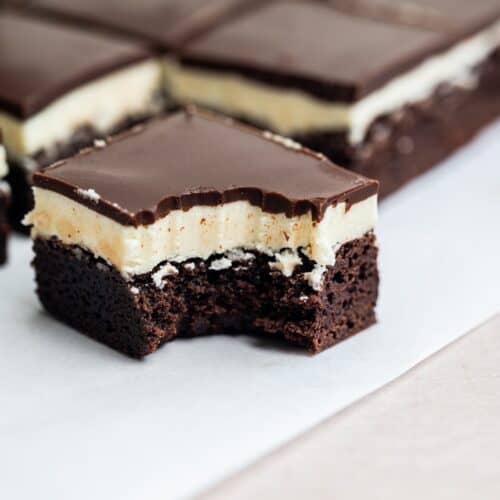 A close-up of a chocolate brownie bar with a creamy filling and chocolate topping, showing a bite taken out of the front piece. Other whole bars are in the background on parchment paper.