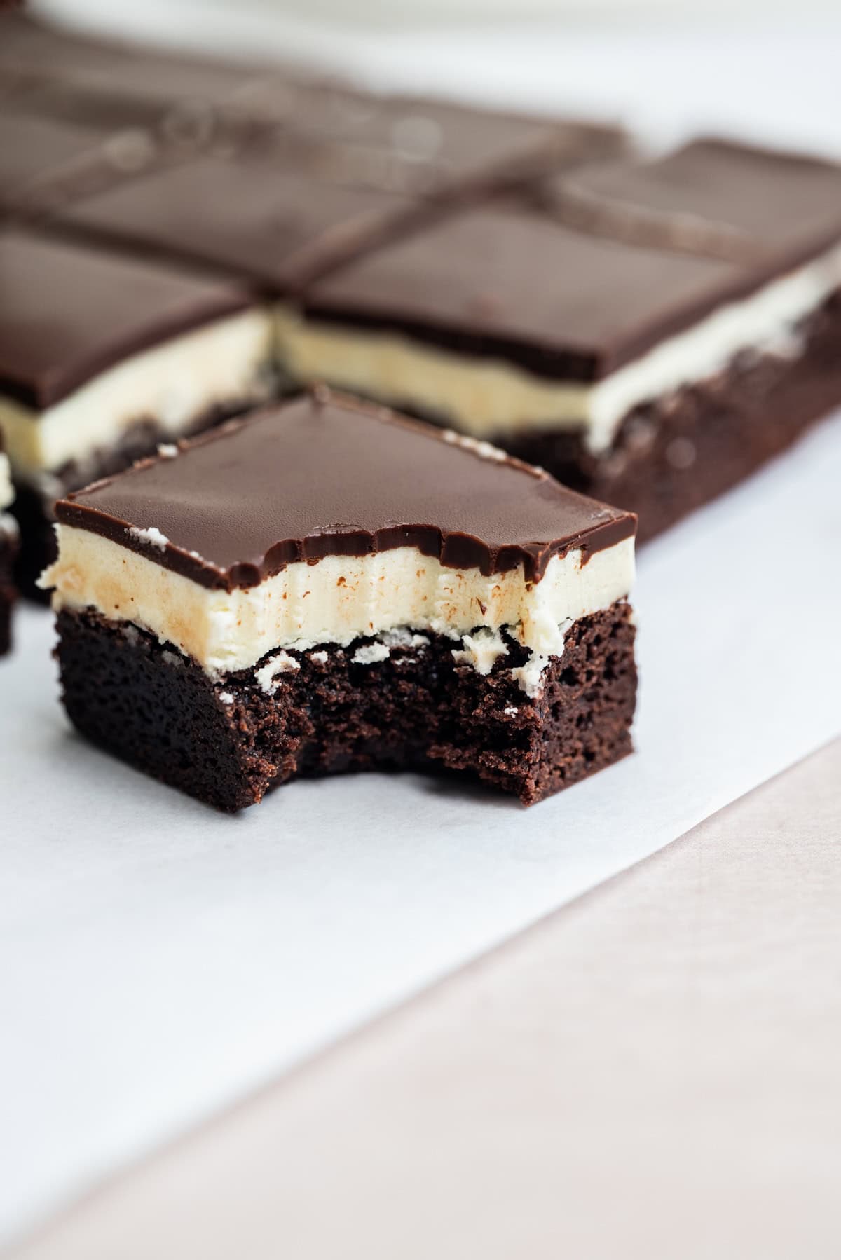 A close-up of a chocolate brownie bar with a creamy filling and chocolate topping, showing a bite taken out of the front piece. Other whole bars are in the background on parchment paper.