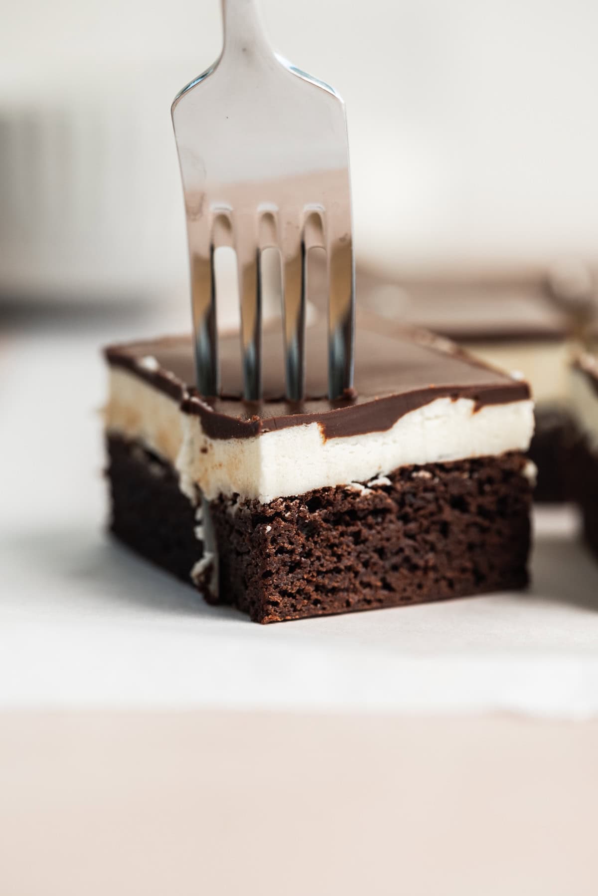 A fork pressed into a layered dessert bar with a brownie base, a creamy white middle layer, and a glossy chocolate top, set on white parchment paper.