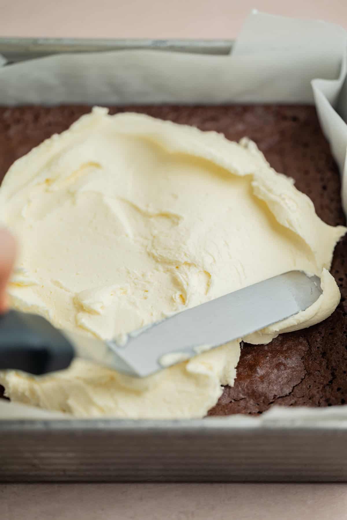 A close-up of a hand using a spatula to spread creamy frosting over a rectangular chocolate cake in a parchment-lined baking pan.
