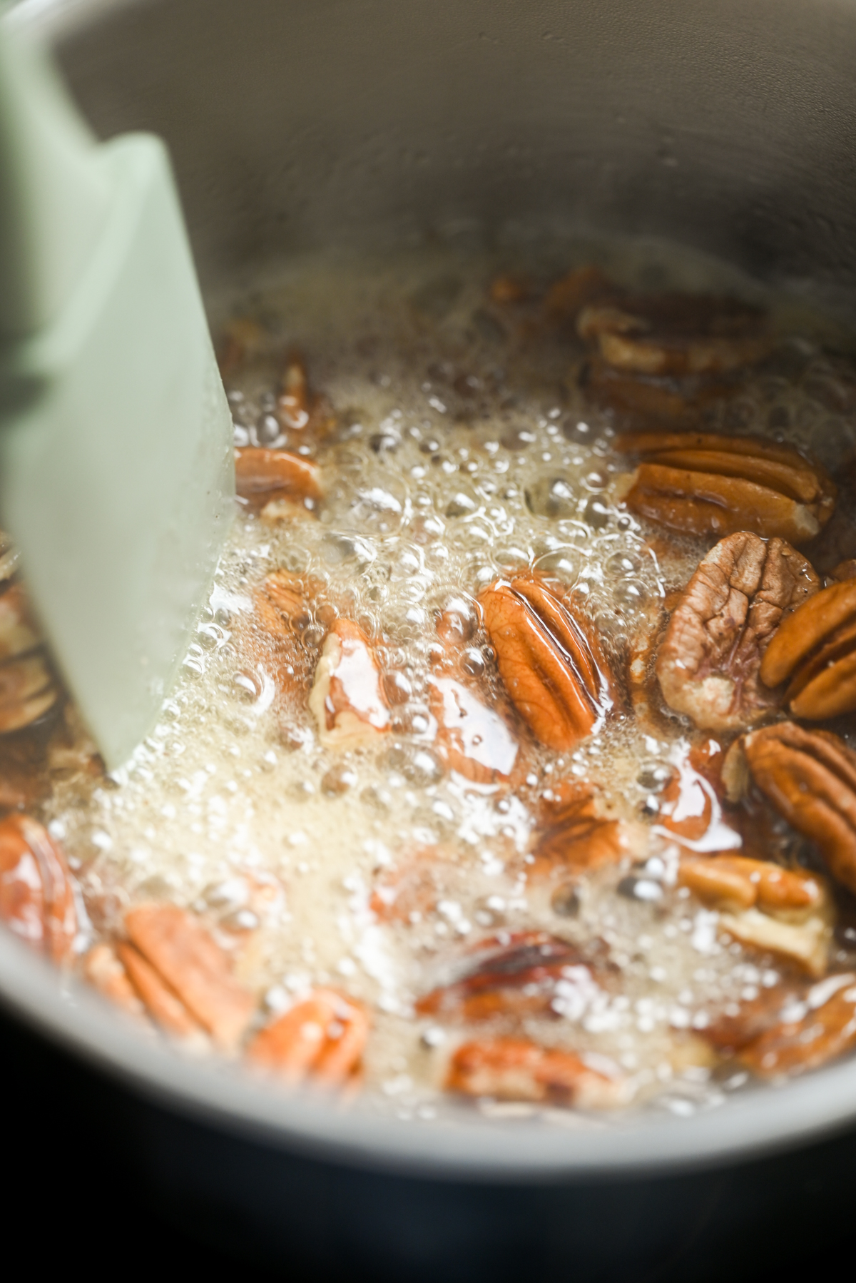 Pecan halves simmering in bubbling sugar syrup in a saucepan, with a spatula stirring the mixture.