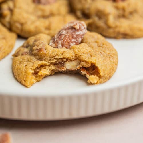 A close-up of a plate with cookies topped with pecans, one cookie in front has a bite taken out, revealing its soft interior.