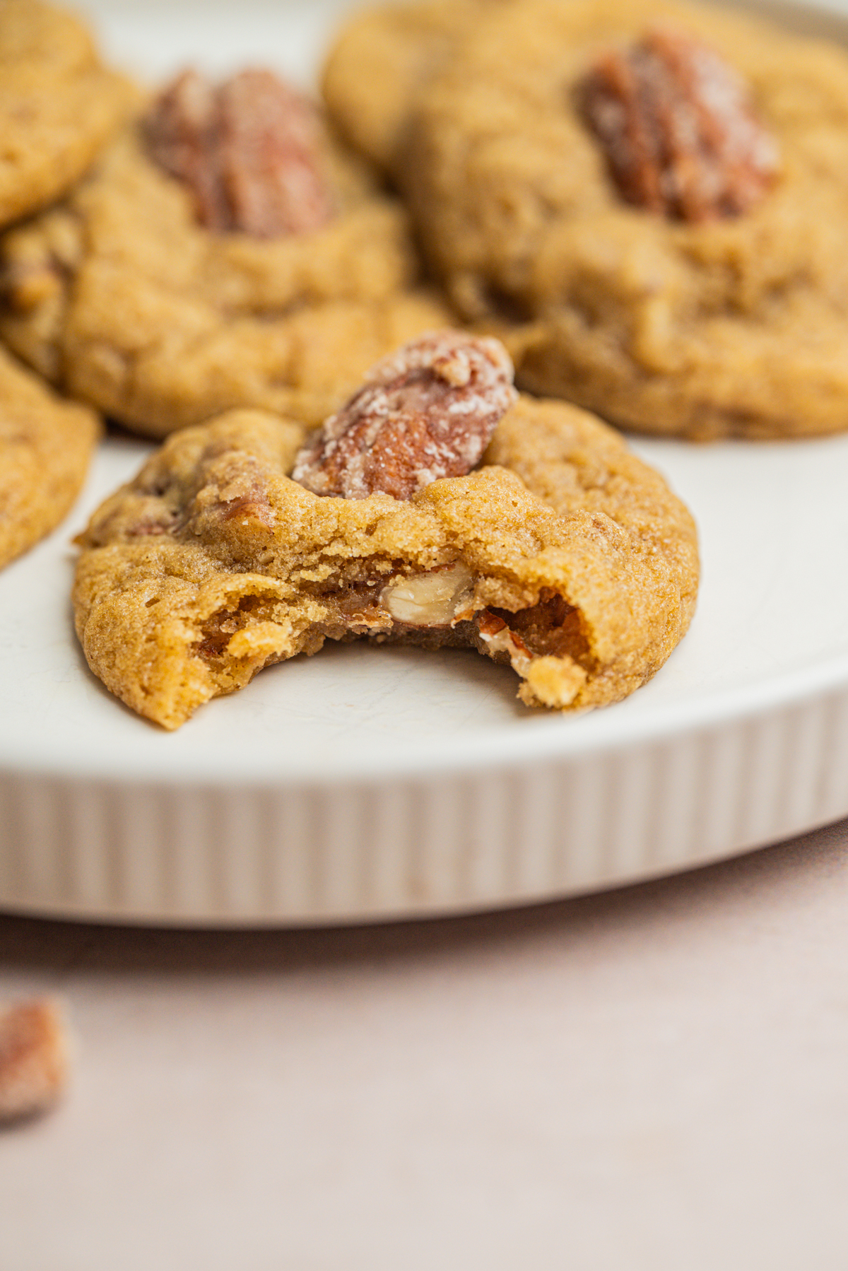 A close-up of a plate with cookies topped with pecans, one cookie in front has a bite taken out, revealing its soft interior.