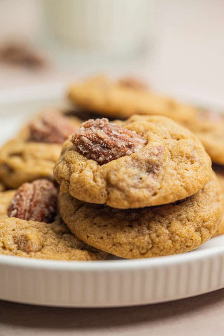 A close-up of a stack of soft brown cookies topped with pecan halves, arranged on a white plate. A blurred glass of milk is visible in the background.
