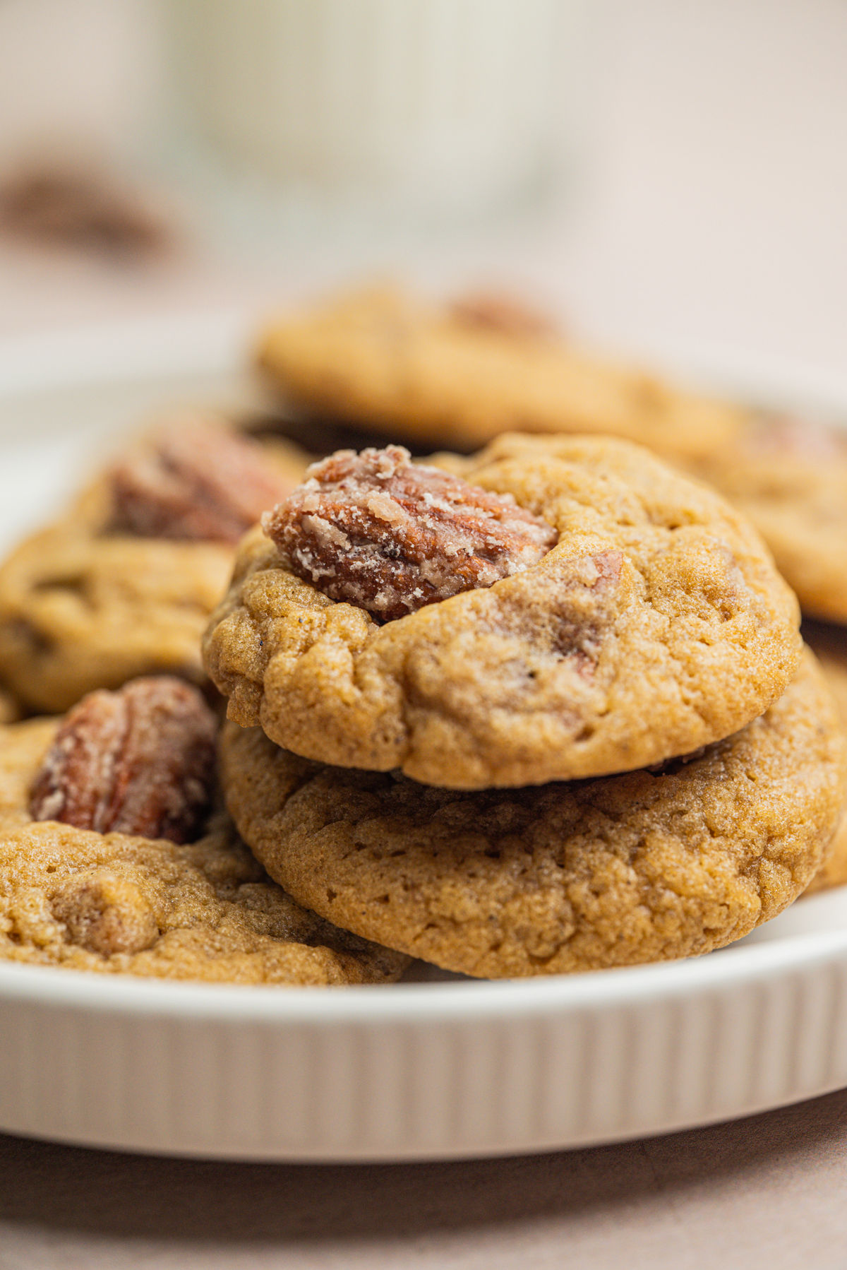 A close-up of a stack of soft brown cookies topped with pecan halves, arranged on a white plate. A blurred glass of milk is visible in the background.