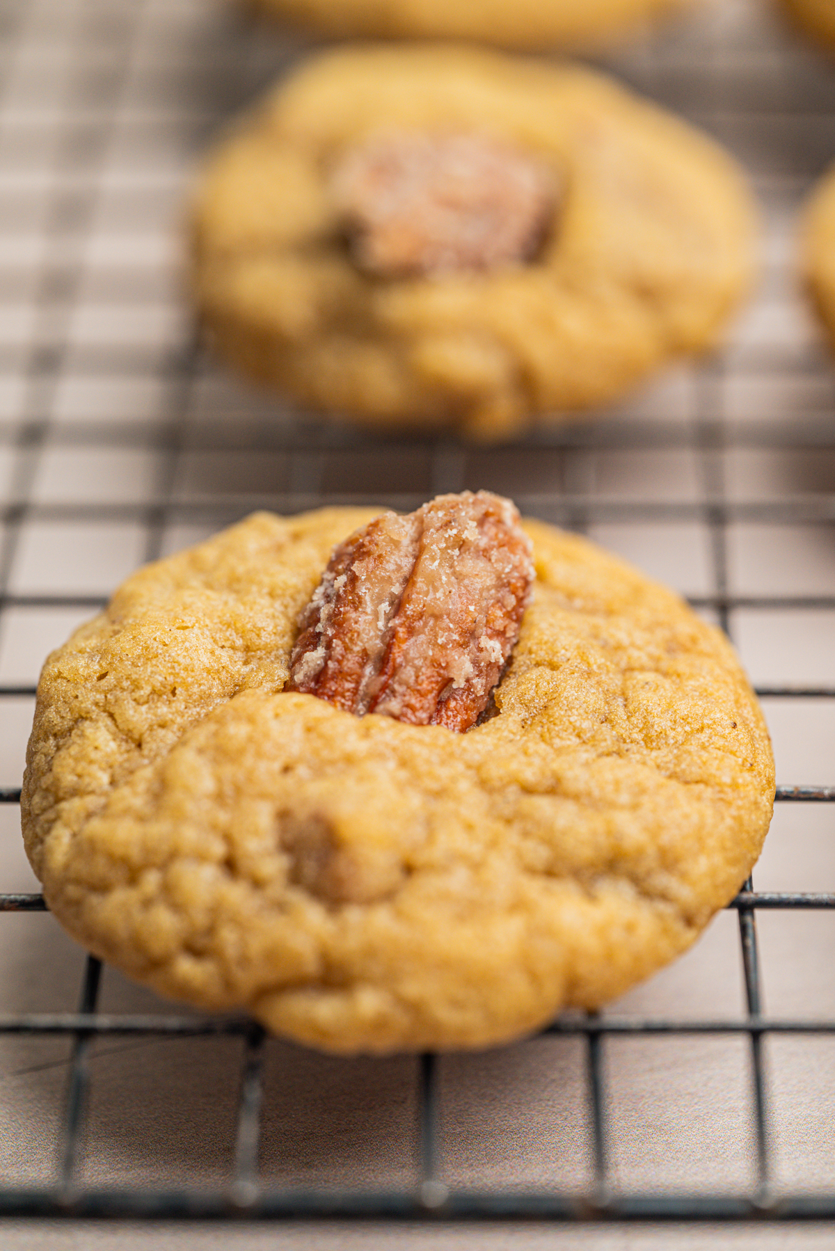 A close-up of a round, golden-brown cookie topped with a pecan half, resting on a black wire cooling rack. Other similar cookies are visible blurred in the background.