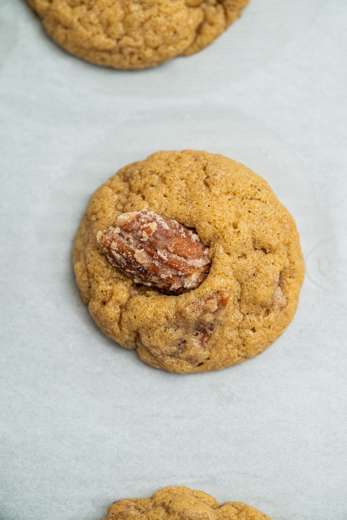 A close-up of a round, golden-brown cookie with a sugared pecan half pressed into the center, placed on a sheet of white parchment paper.