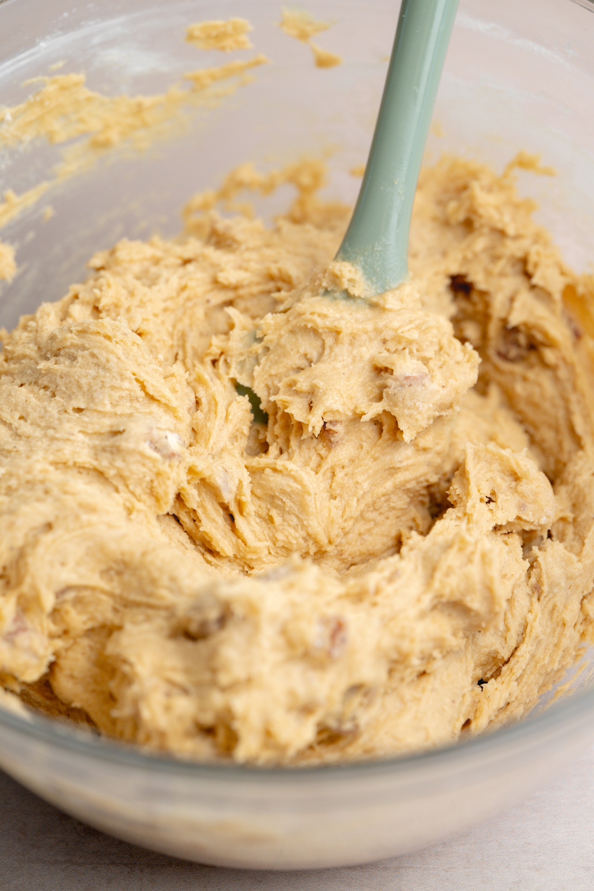 A glass bowl filled with thick cookie dough being mixed with a green spatula.