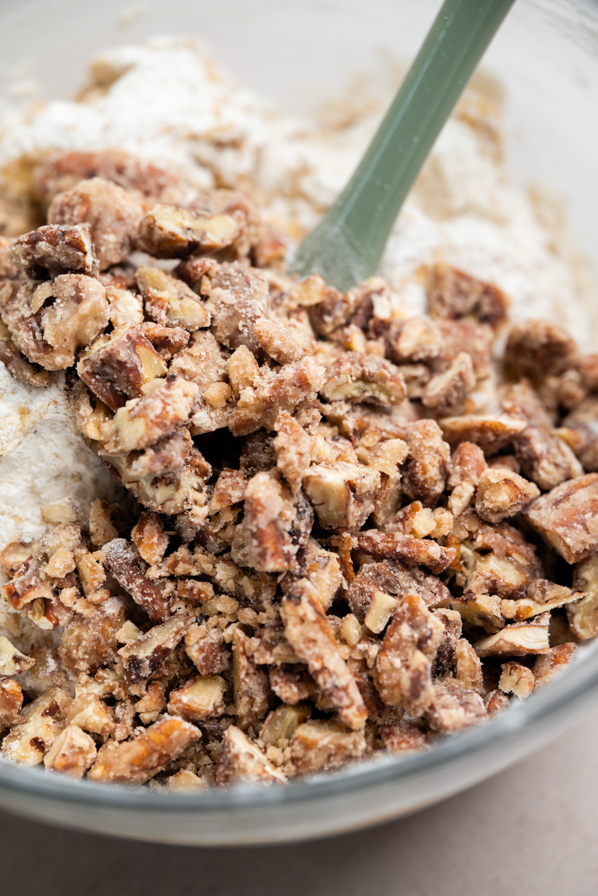Chopped pecans coated in flour and sugar mixture sit in a glass mixing bowl with a green spatula, ready to be mixed into a baking batter.