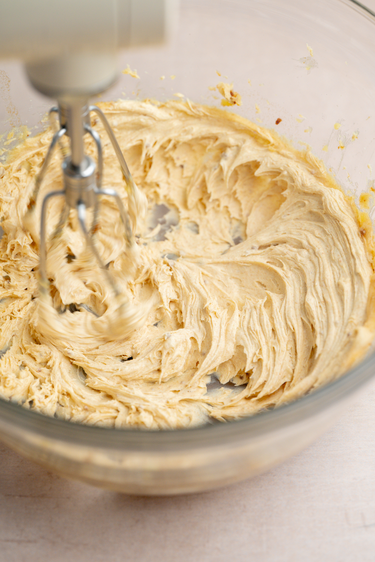 An electric hand mixer is blending creamy, light-colored batter in a clear glass bowl on a light surface.