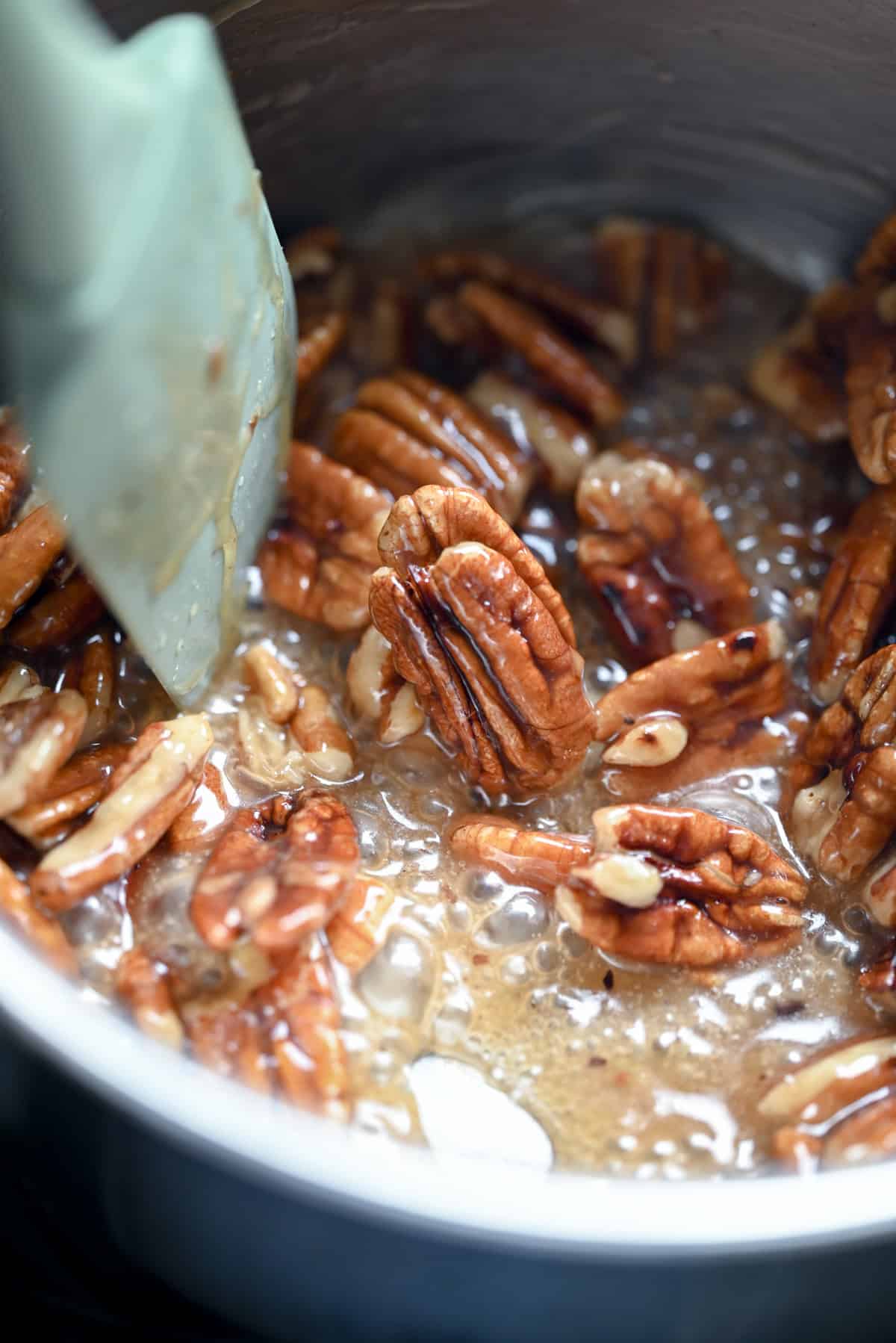 Pecans being stirred with a spatula in a saucepan filled with bubbling caramelized sugar or syrup.