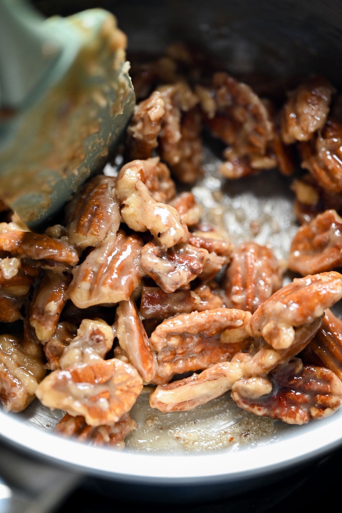 Close-up of glazed pecans being stirred with a spatula in a metal pan, with a glossy, caramelized coating visible on the nuts.