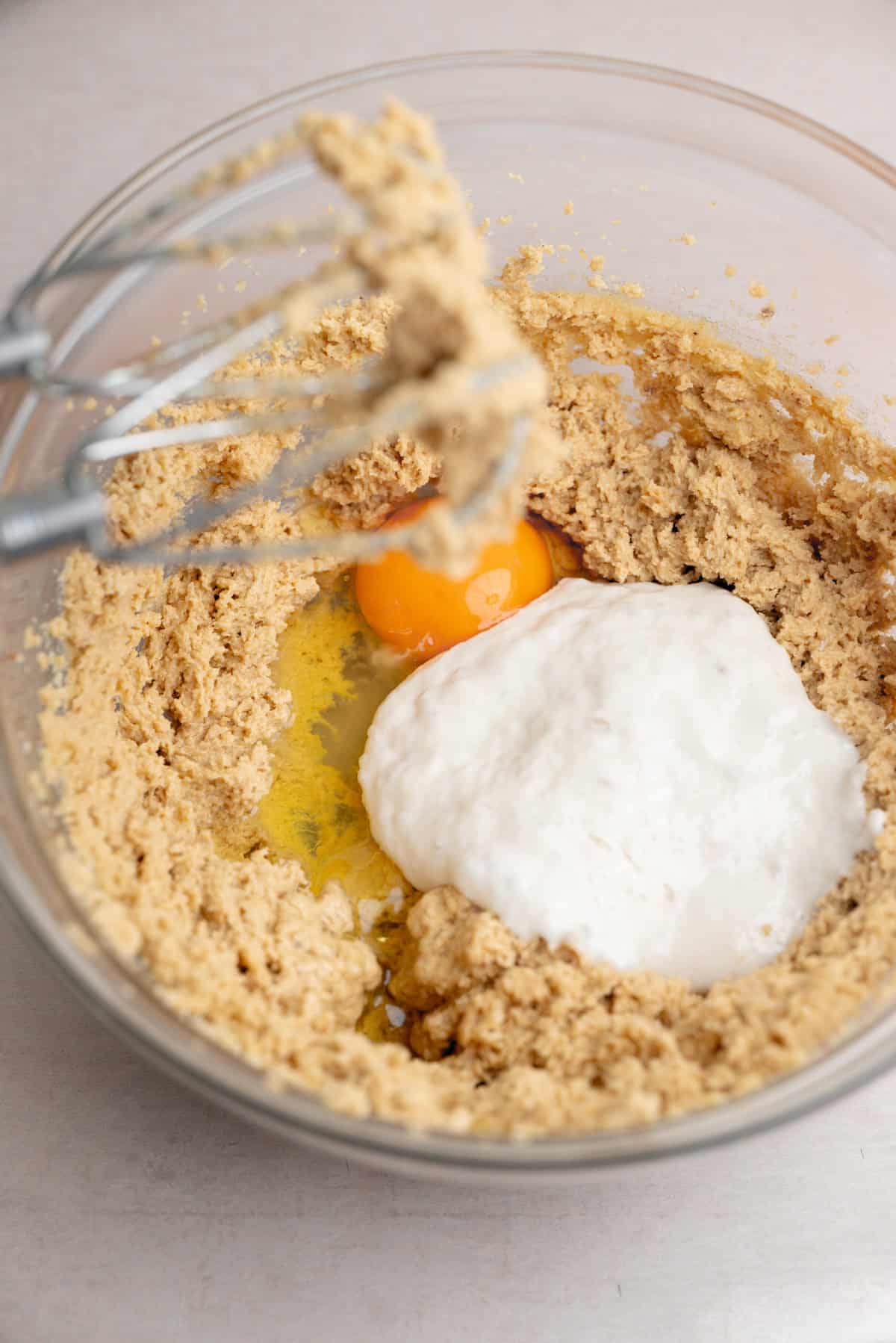 A glass bowl with a mixture of creamed sugar and butter, an egg, sourdough discard starter and a metal hand mixer resting on the edge.