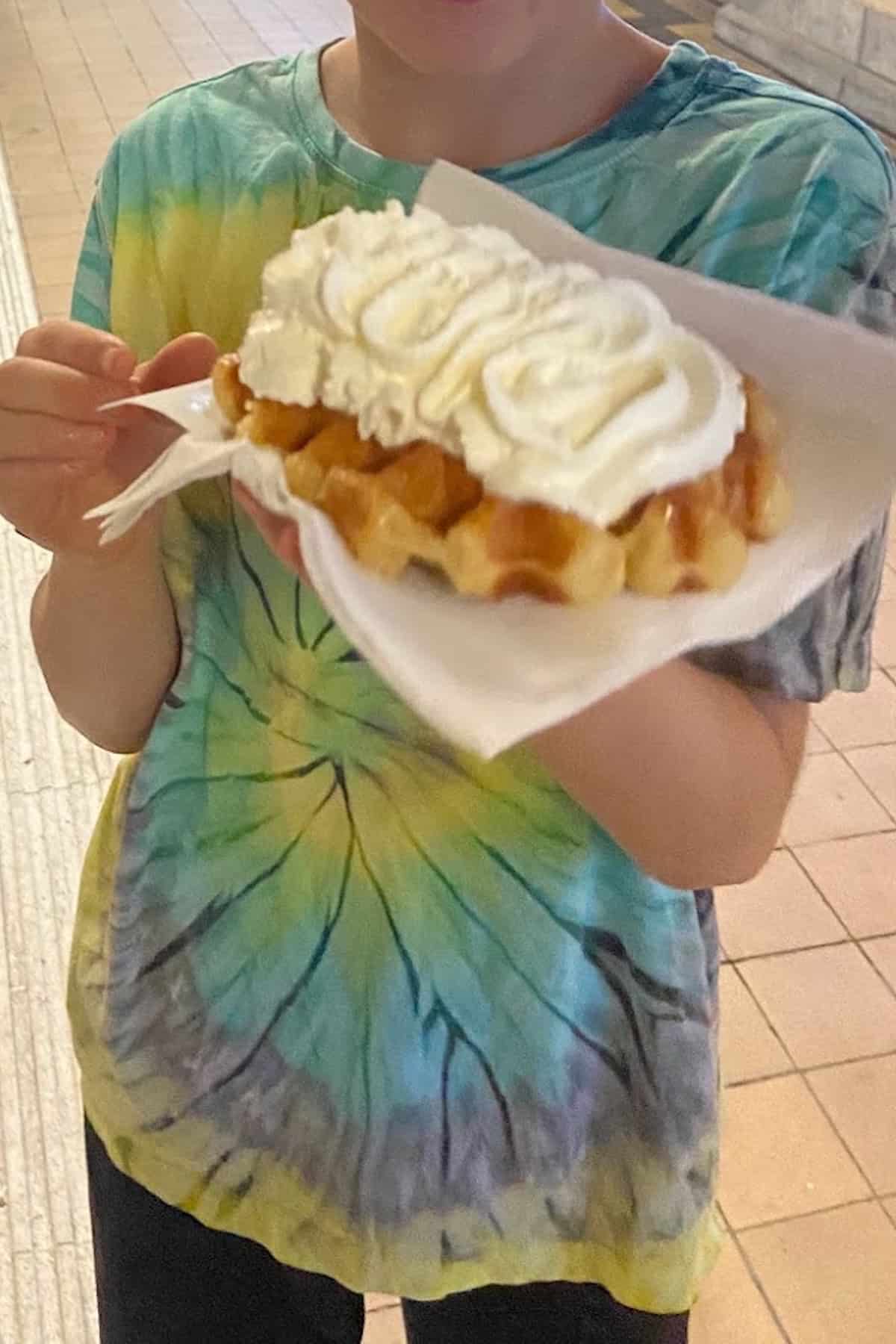 A person wearing a tie-dye shirt holds a Liège waffle topped with whipped cream on a napkin, using a fork.