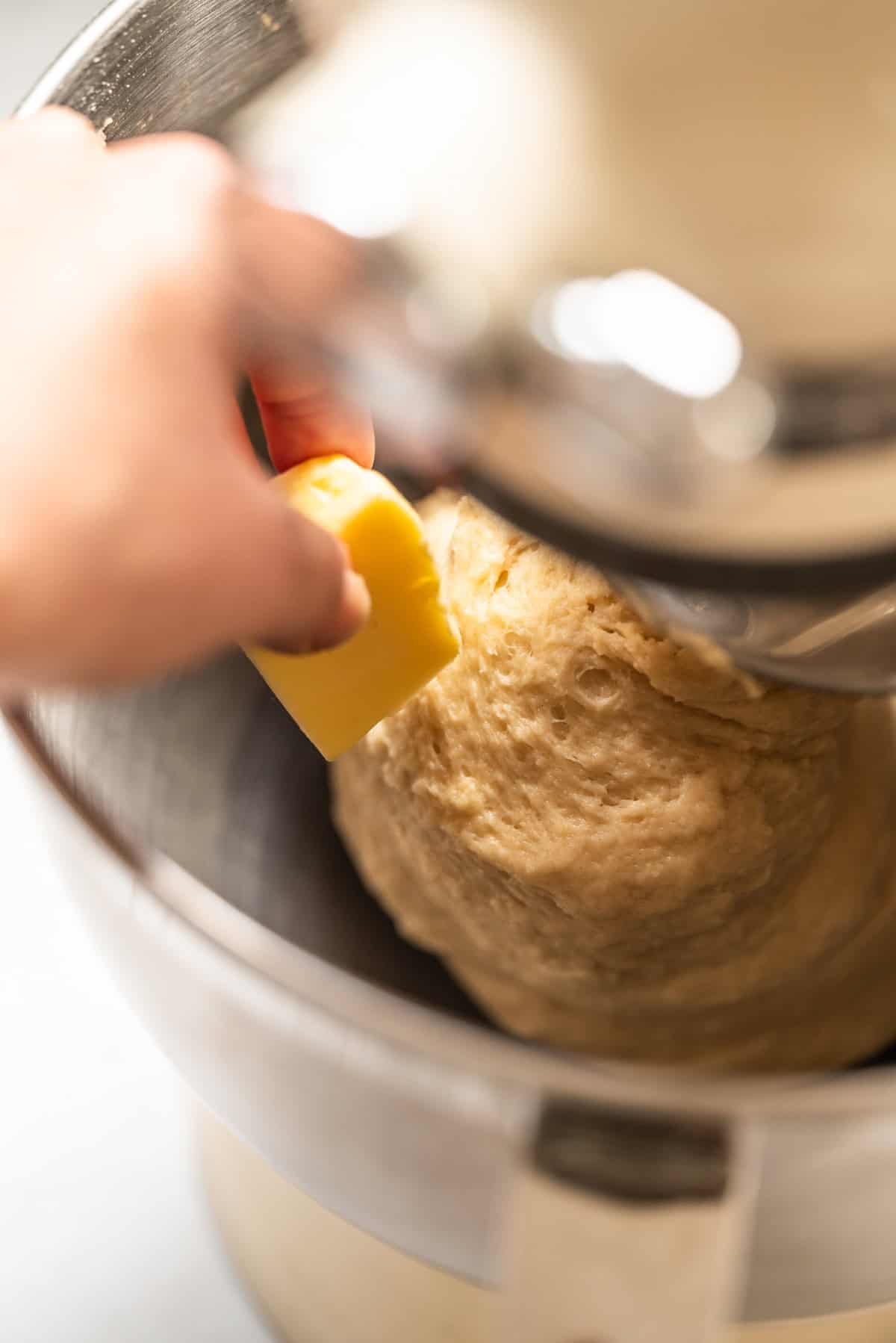 A hand adds a chunk of butter to dough mixing in a stand mixer.
