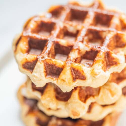 A close-up of three stacked golden-brown leige waffles on a white plate, with a light dusting of sugar visible on top.