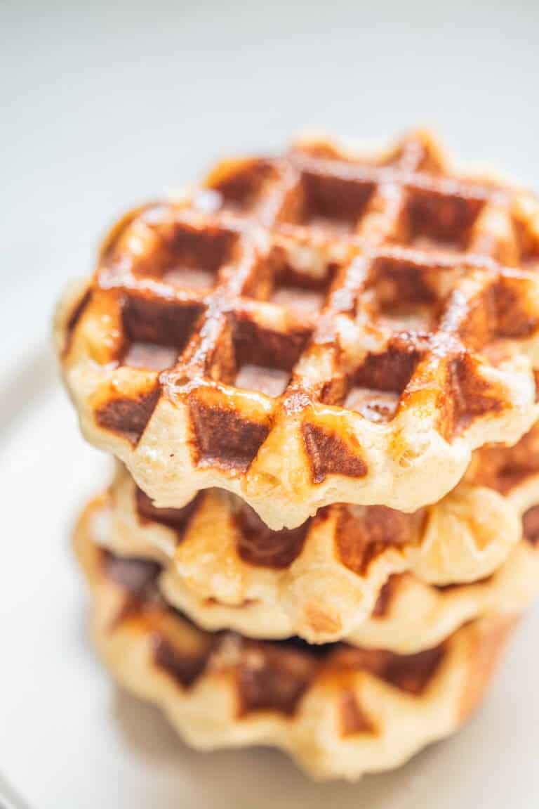 A close-up of three stacked golden-brown leige waffles on a white plate, with a light dusting of sugar visible on top.